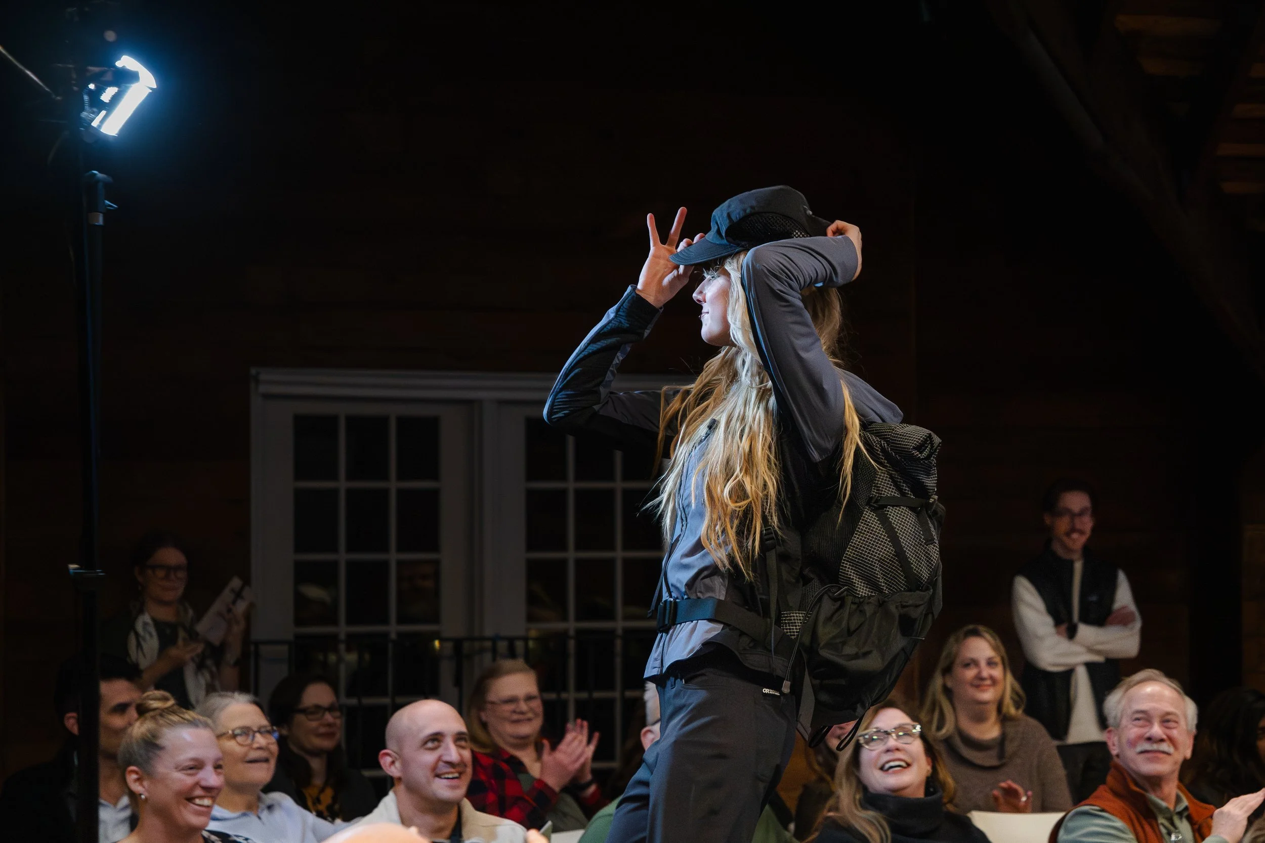 A woman wearing outdoor gear and a backpack, holding a hat, standing in front of an audience that is laughing and clapping.