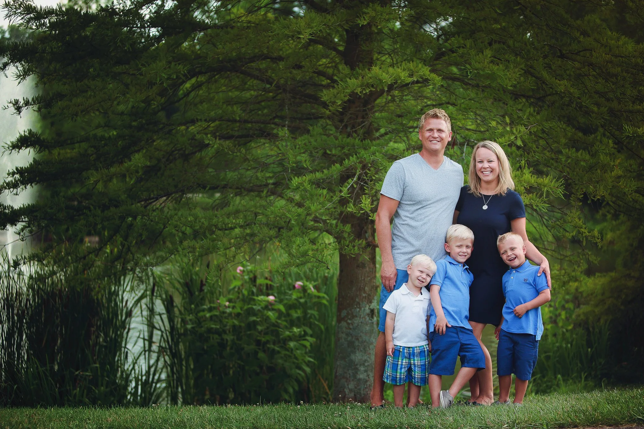A family of five, including a man, woman, and three young boys, standing outdoors in front of a large green tree, smiling at the camera.
