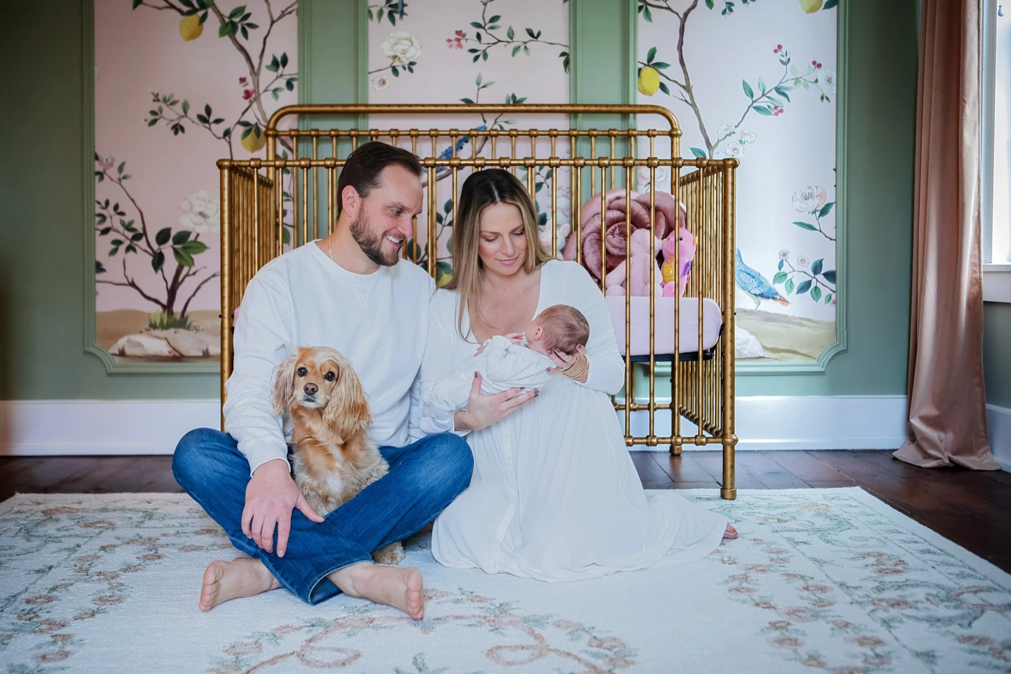 A family sitting on the floor of a nursery, holding a newborn baby. The man has a dog on his lap, and they are all looking at the baby affectionately. The room has a mural on the wall behind them and a golden crib in the background.