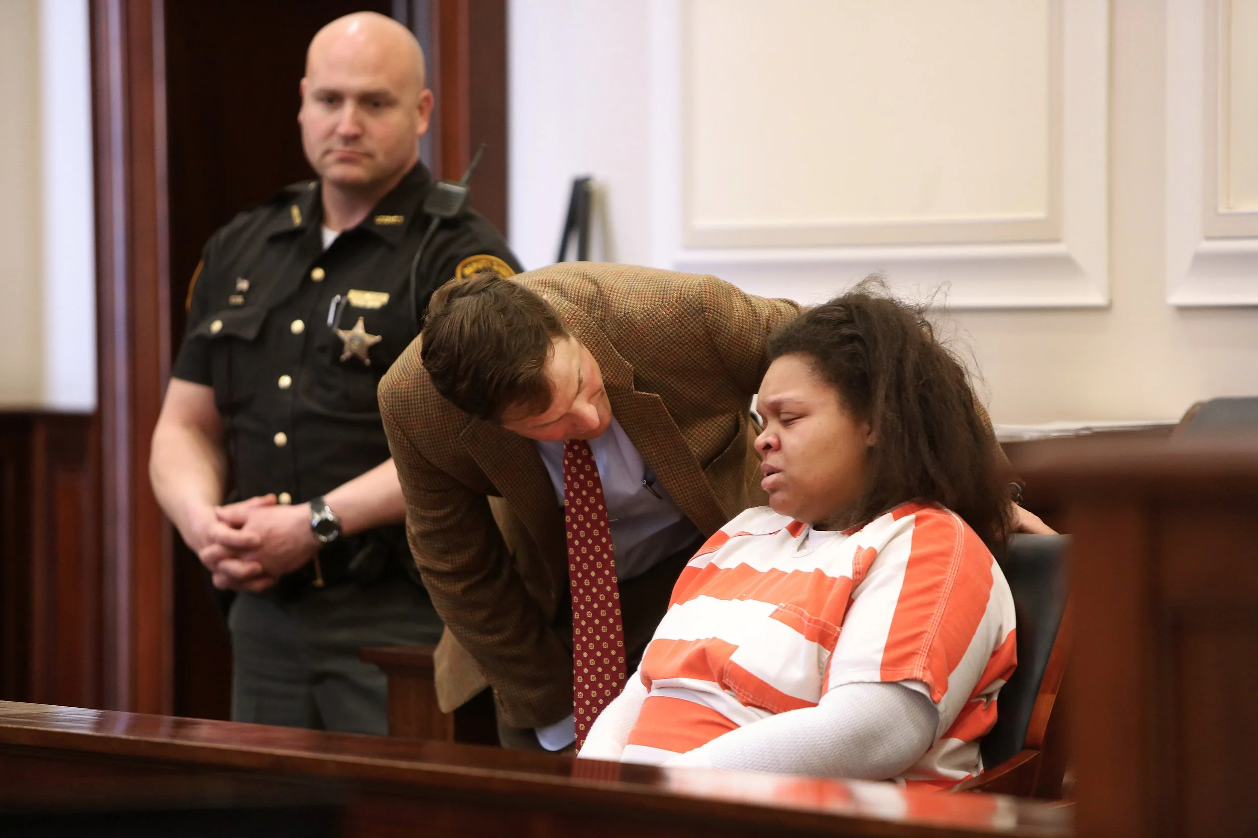 A woman in an orange and white striped prison jumpsuit sitting in a courtroom, crying and leaning on a chair. A man in a brown blazer and red patterned tie leans over her, appearing to comfort her. A police officer in uniform stands in the background