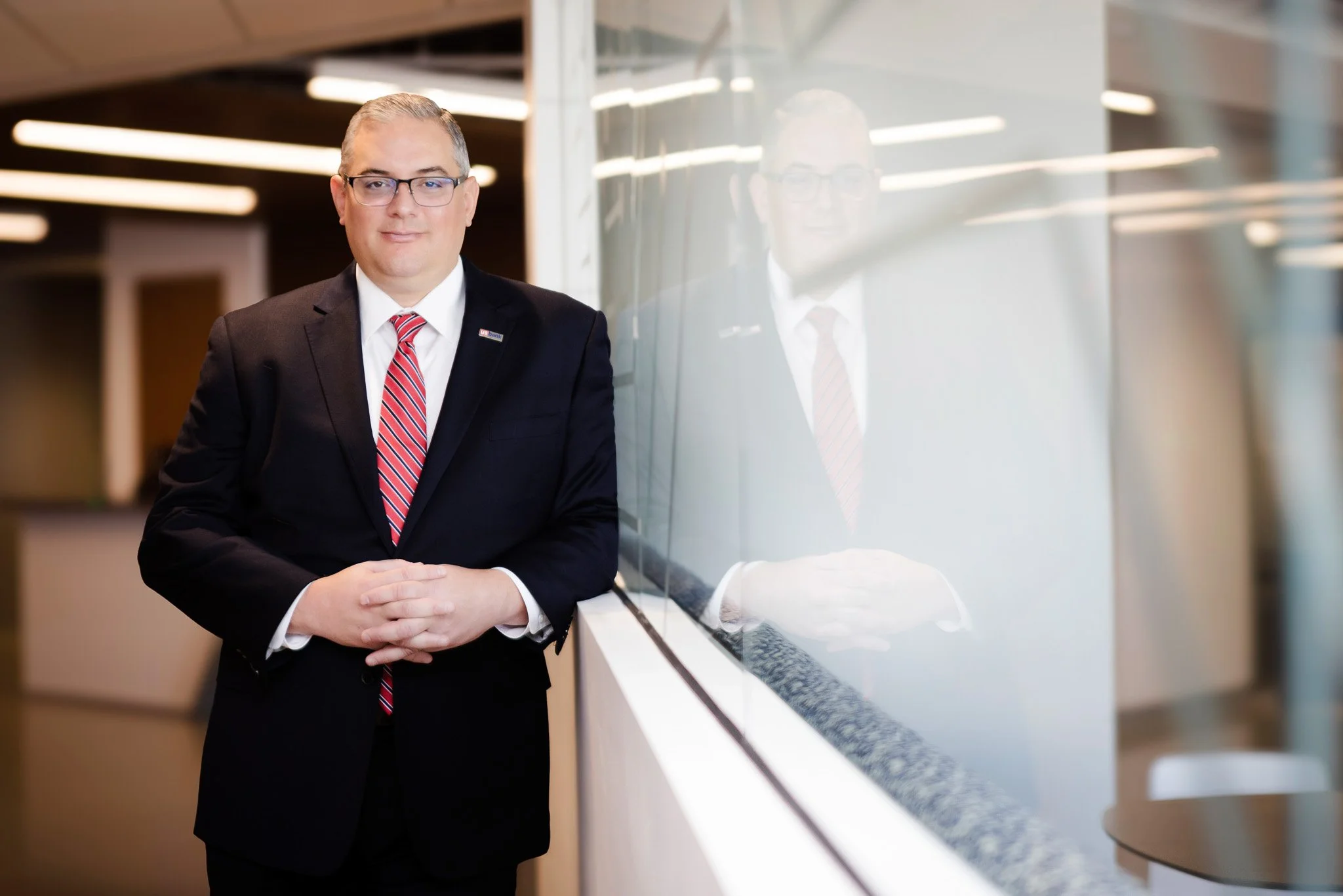 A man in a suit and tie standing next to a glass wall, with his reflection visible in the glass.