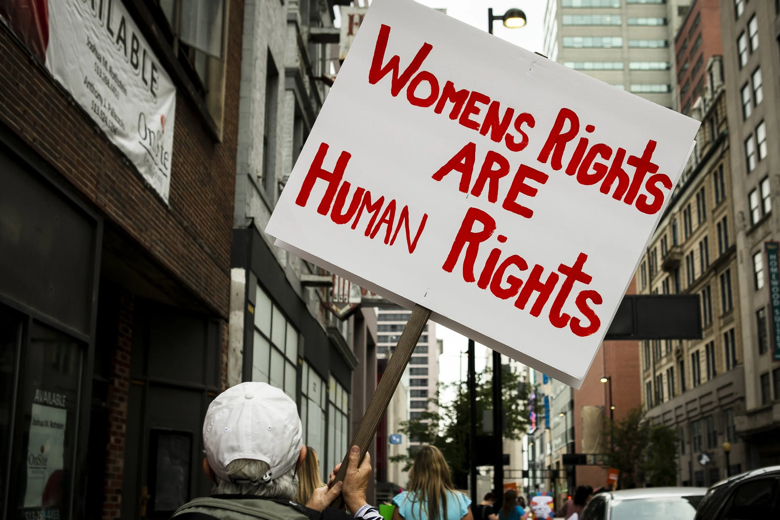 A person wearing a white cap holding a sign that says, 'Women’s Rights Are Human Rights,' during a street protest in an urban area.