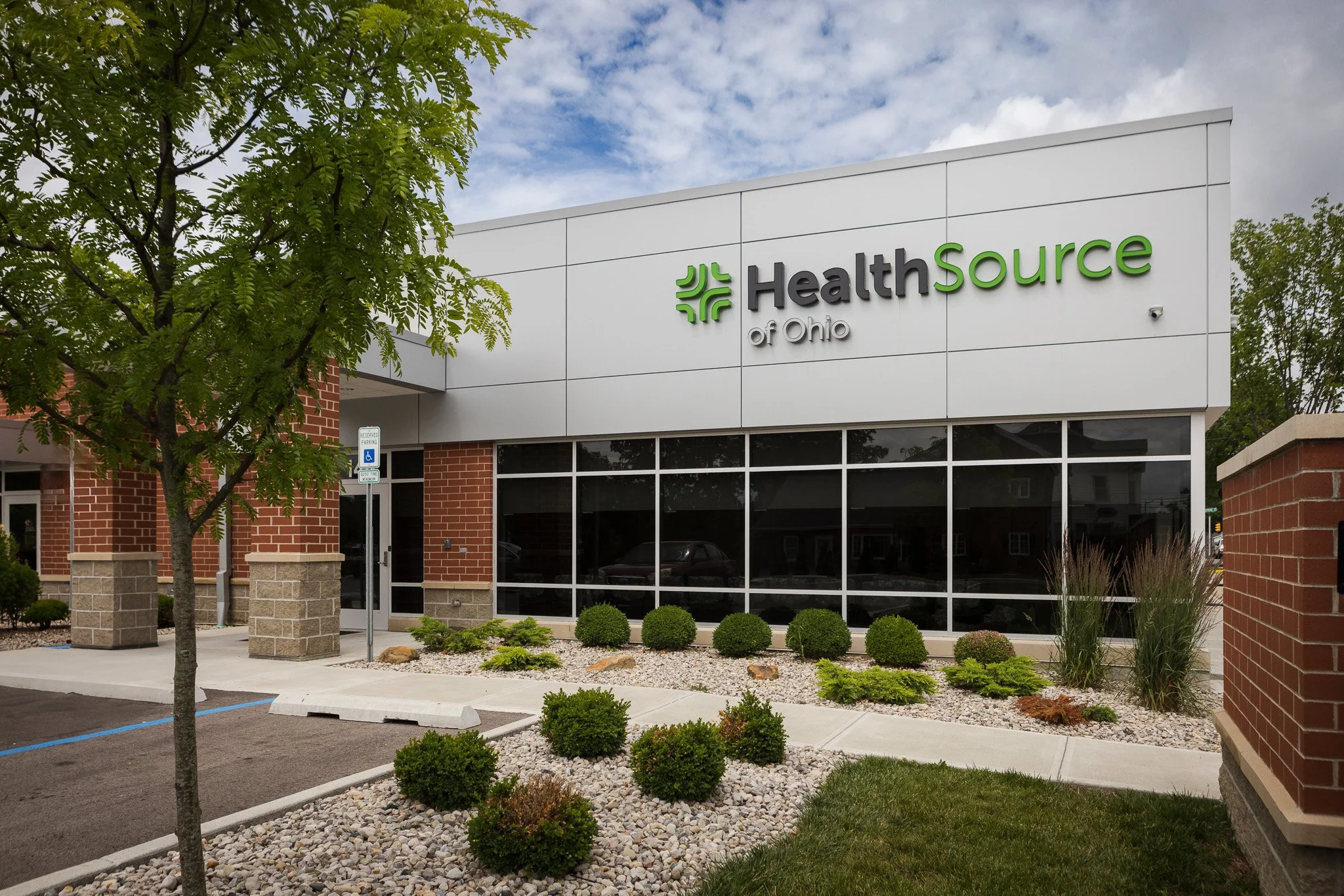 Exterior view of a modern building with a sign that reads 'HealthSource of Ohio,' surrounded by landscaping with bushes, a small tree, and rocks, under a partly cloudy sky.