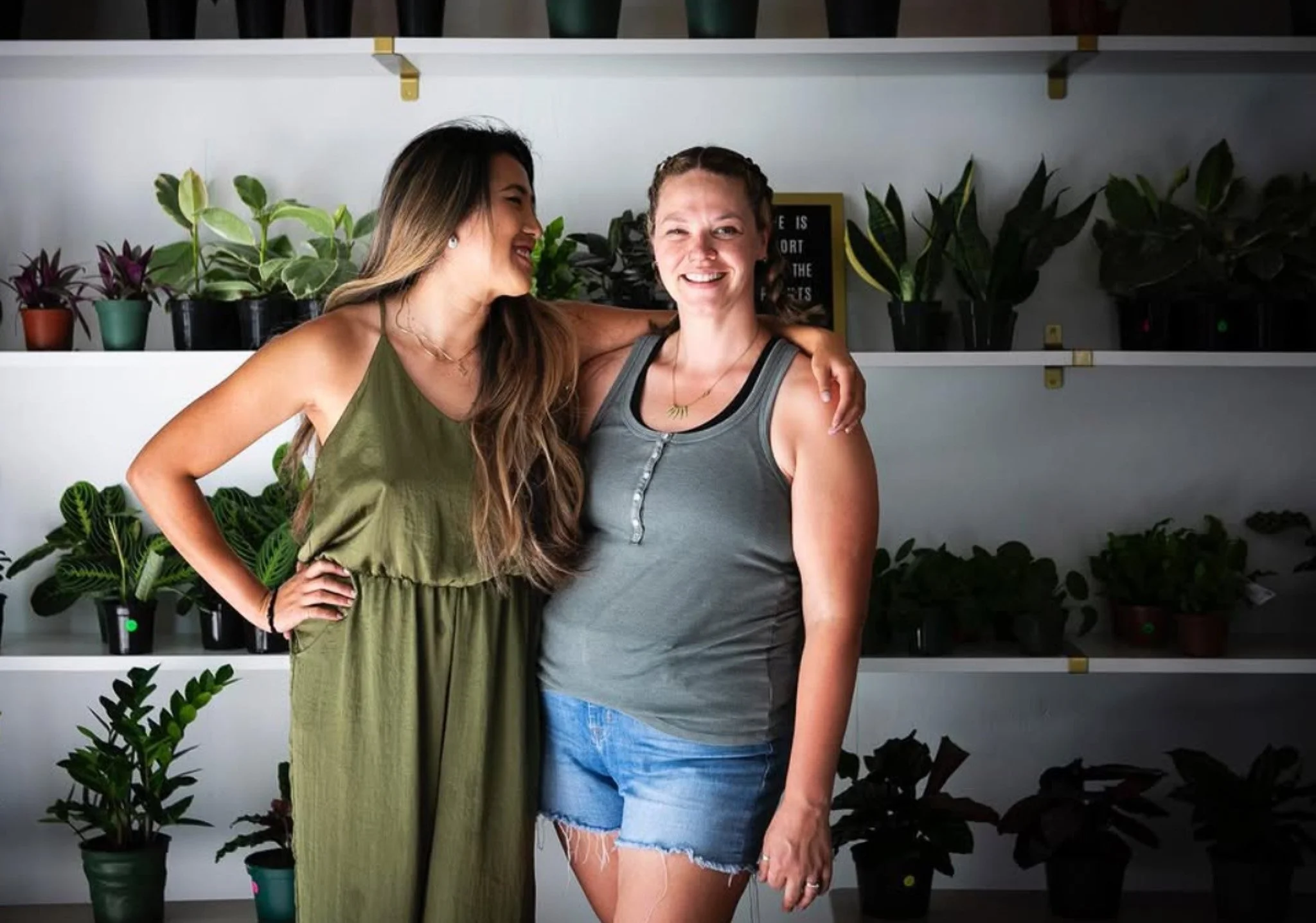 Two women smiling and hugging in front of shelves filled with potted plants.