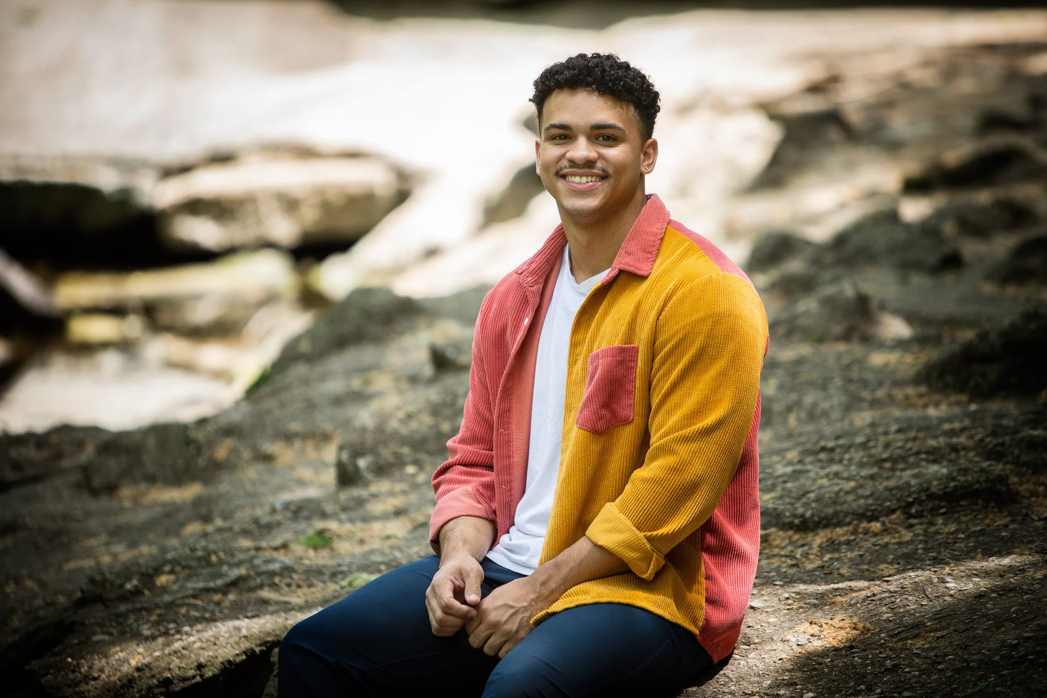 A smiling young man sitting on rocks near a body of water.
