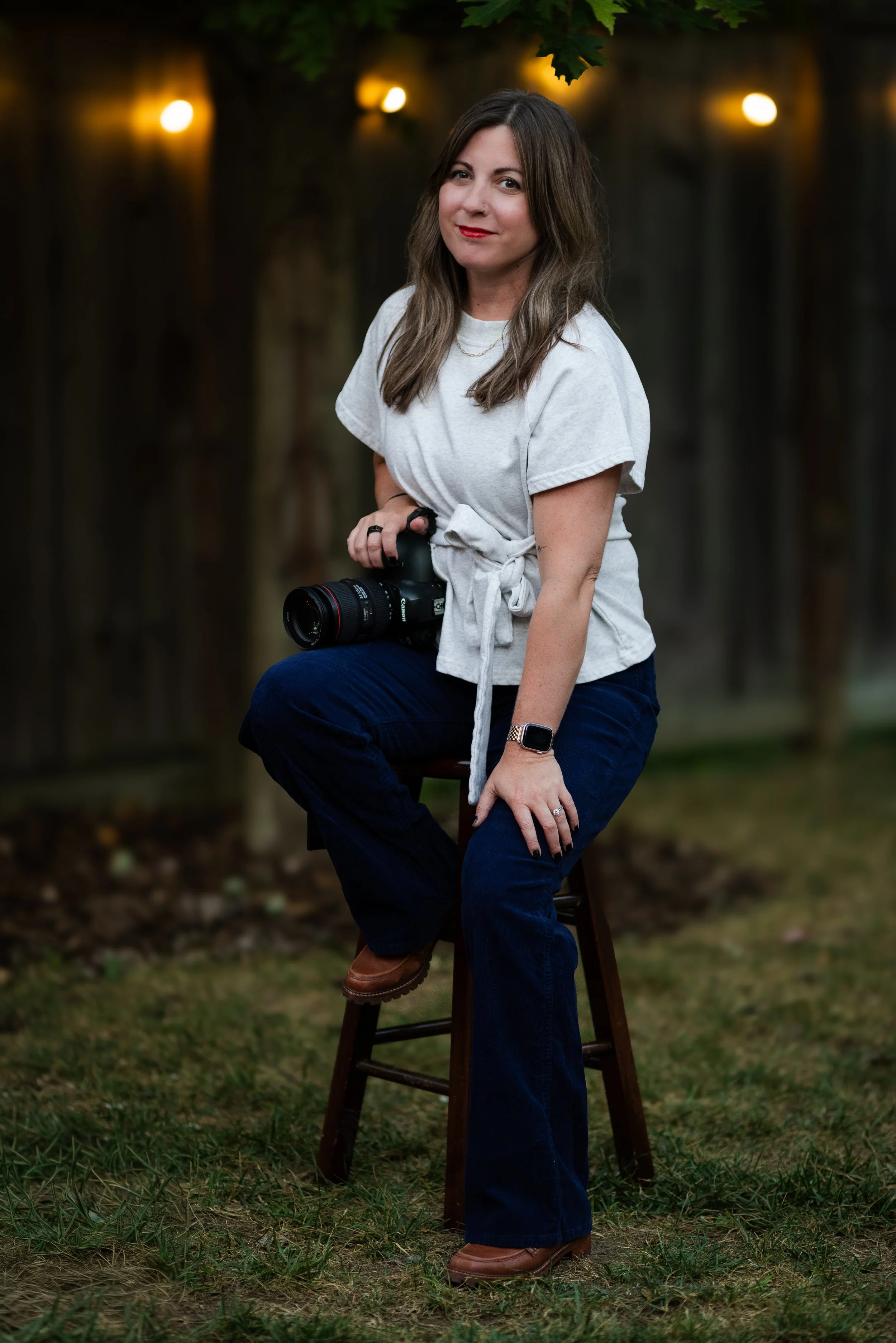 A woman sitting on a wooden stool outdoors, holding a professional camera, wearing a white tie-front t-shirt, dark blue jeans, brown shoes, and an Apple Watch, with blurred greenery and warm outdoor lighting in the background.