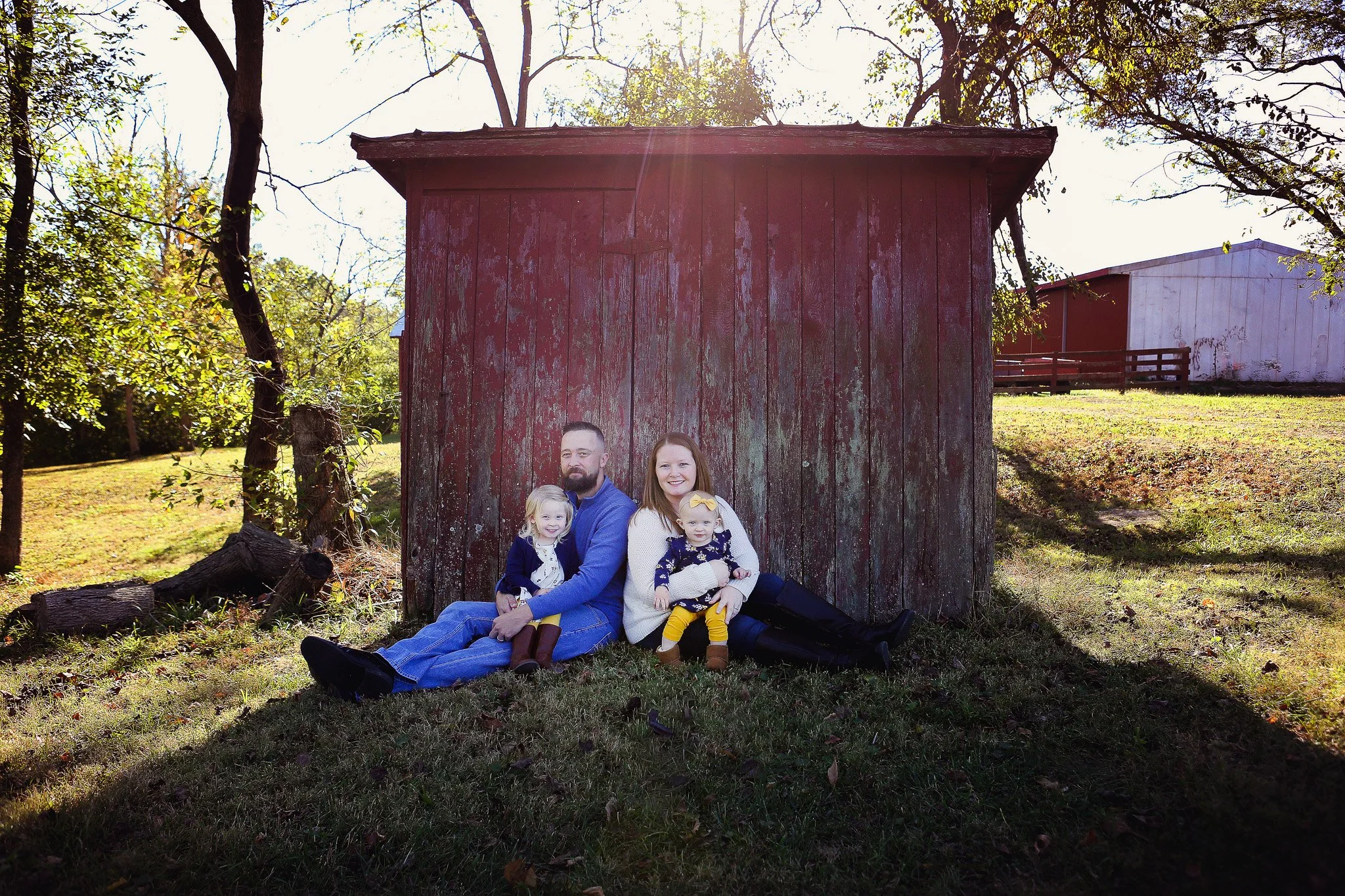 A family of four sitting on the grass in front of a weathered red barn, with trees and a white building in the background, on a sunny day.