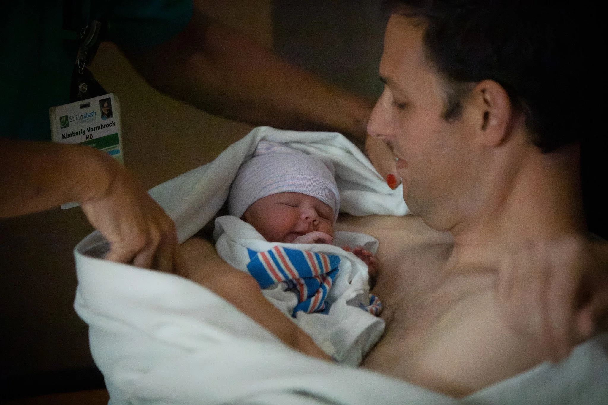 A man with dark hair lying on a bed, smiling and looking at a newborn baby, who is wrapped in a hospital blanket, wearing a hat, and is smiling with eyes closed. The man is shirtless, and the baby is partially covered by a white hospital sheet.