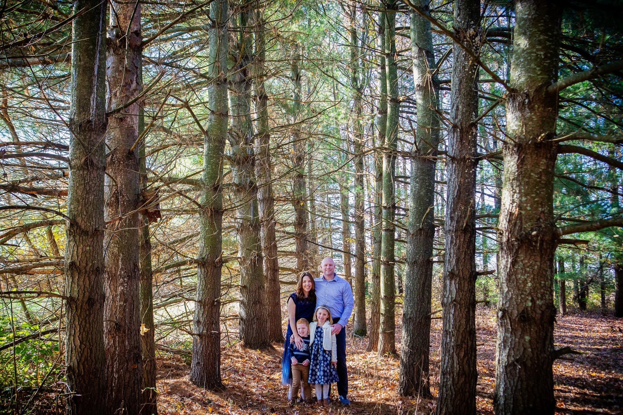 A family of four standing in a wooded forest with tall trees and fallen leaves on the ground, smiling at the camera.