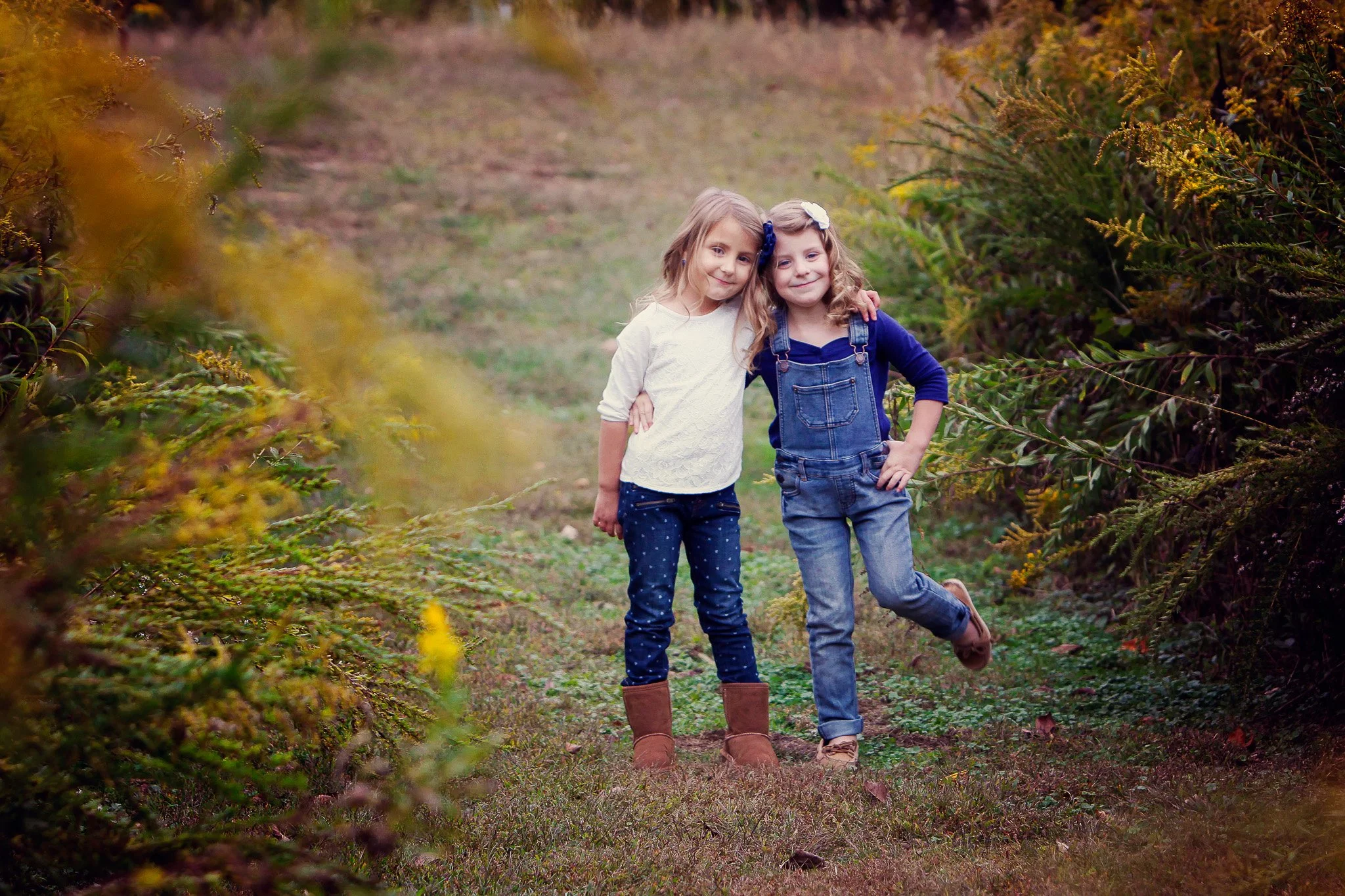Two young girls standing close together on a grassy path surrounded by green foliage, smiling at the camera.