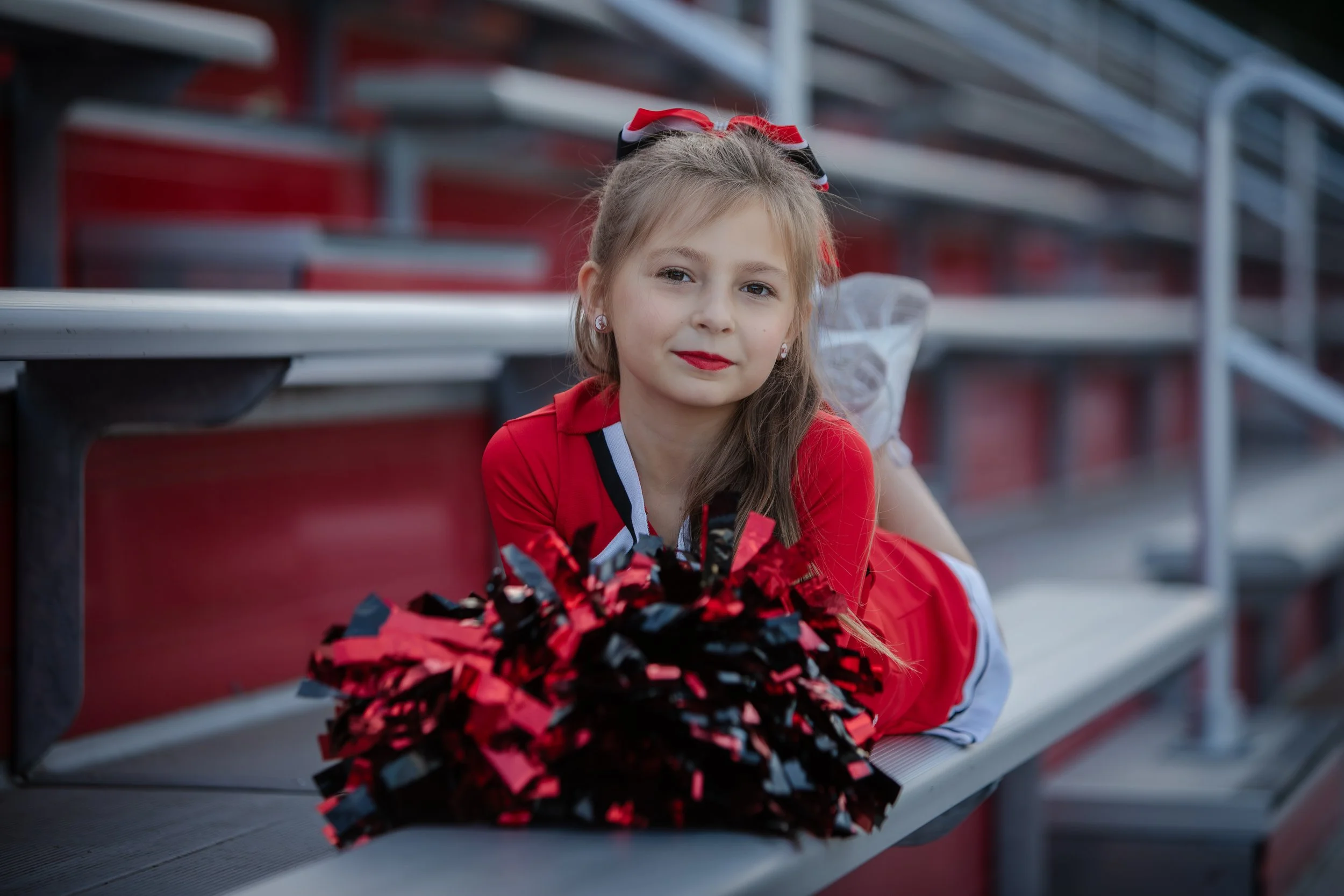 A young girl dressed in a red school cheerleading outfit with a bow in her hair, sitting on stadium bleachers with a black and red pom-pom in front of her.