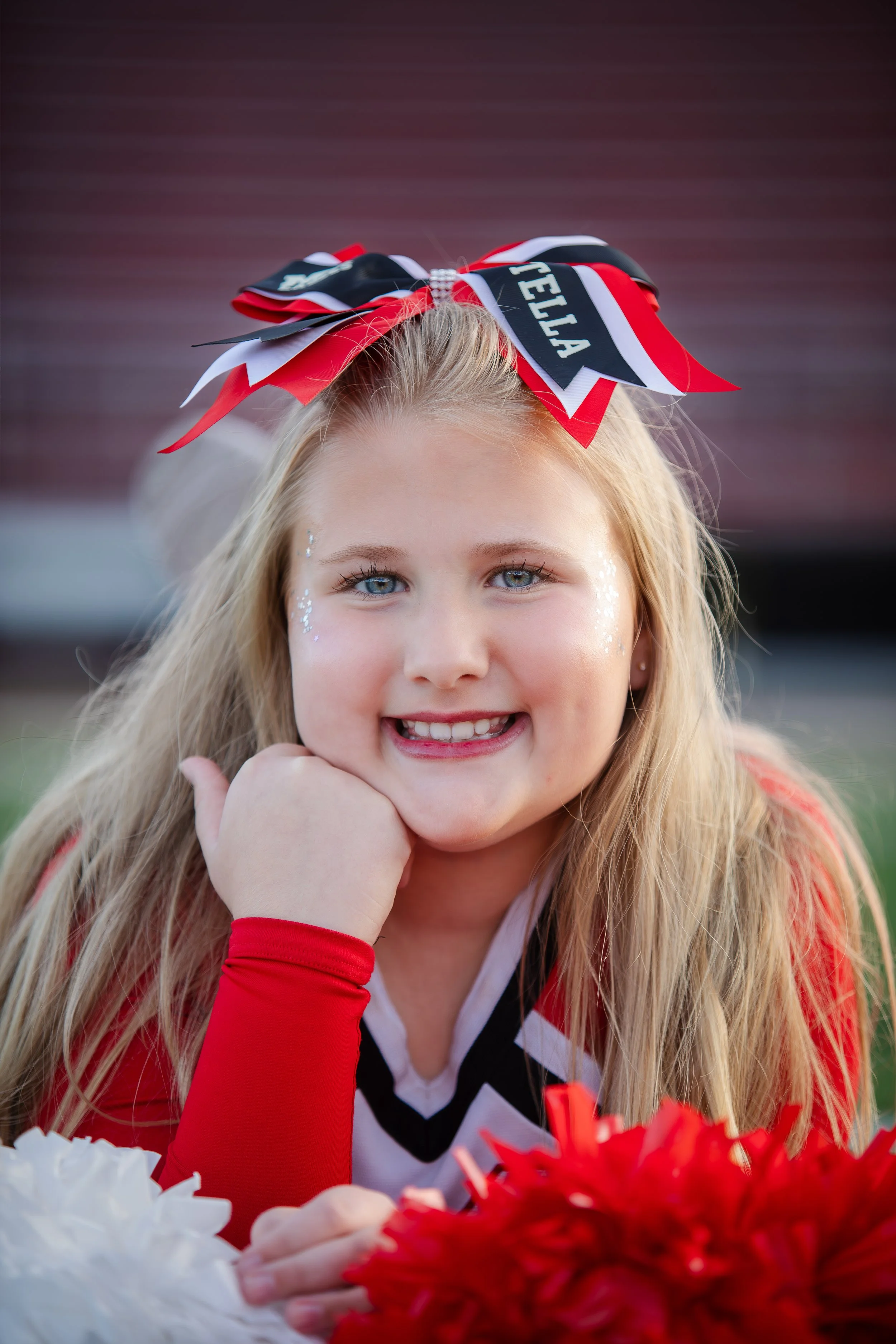 A young girl with long blonde hair, blue eyes, and a big smile, wearing a cheerleader outfit with red and white colors, and a large red, white, and black cheerleading bow on her head. She has sparkles on her cheeks and is lying on her stomach with he