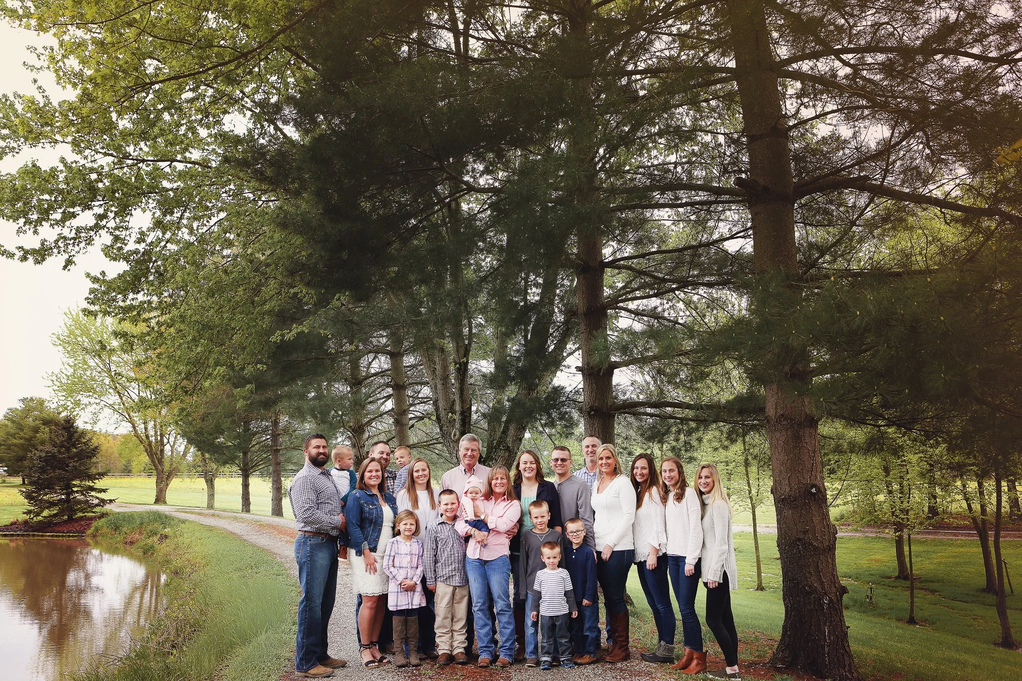 A large family group posing outdoors on a park path with a pond on the left and tall trees in the background.