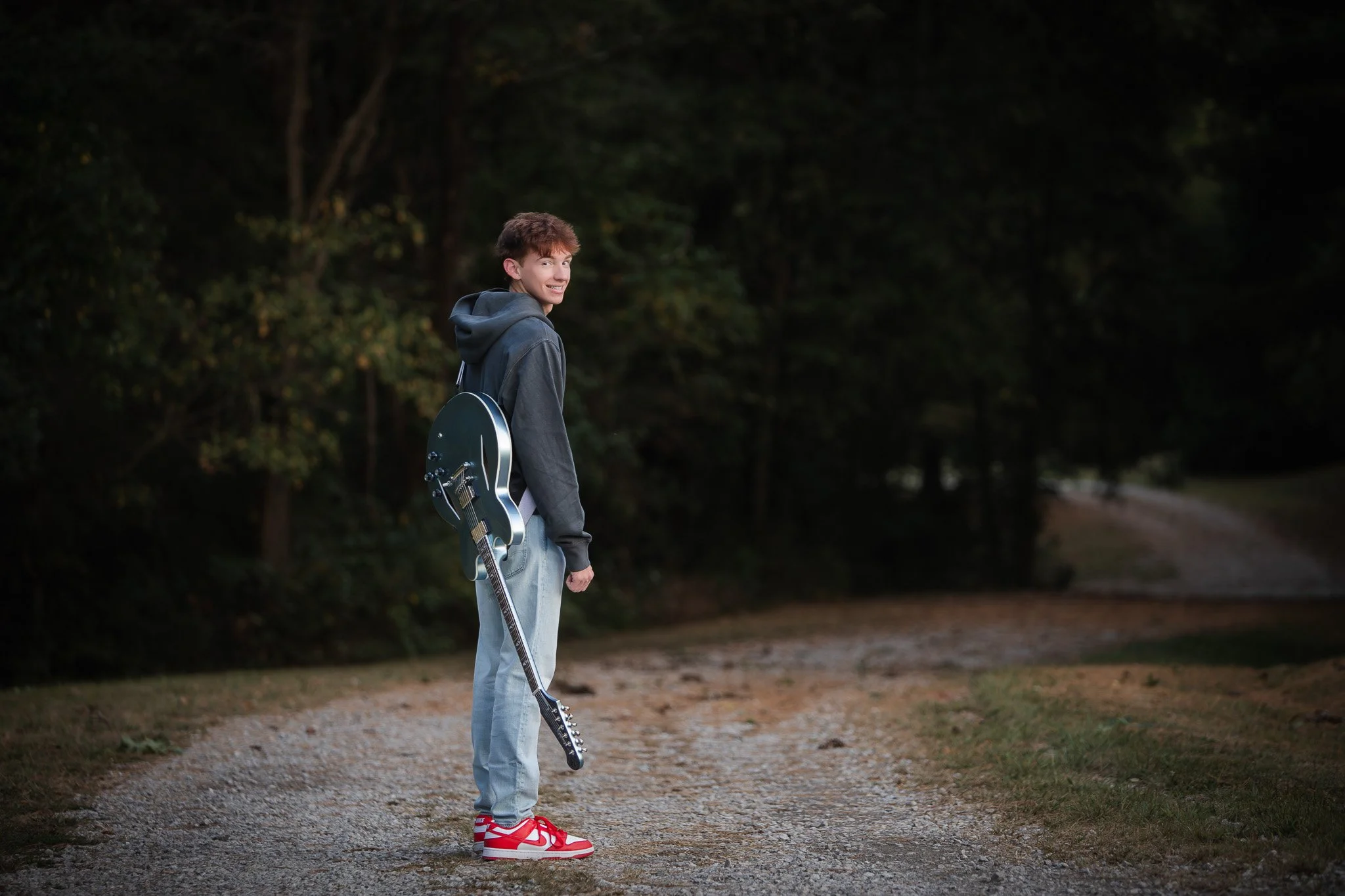 A young man in casual clothing with a hoodie, jeans, and red sneakers stands on a dirt path in a wooded area, carrying a black electric guitar over his shoulder, and looking back over his shoulder smiling.