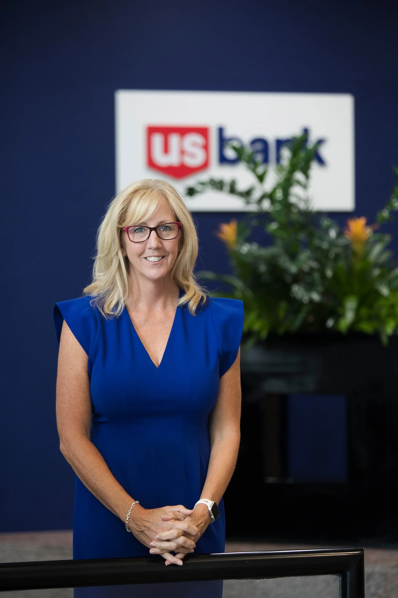 A woman with blond hair, glasses, and a blue dress standing in front of a US Bank sign and a large planter with green leaves and yellow flowers.