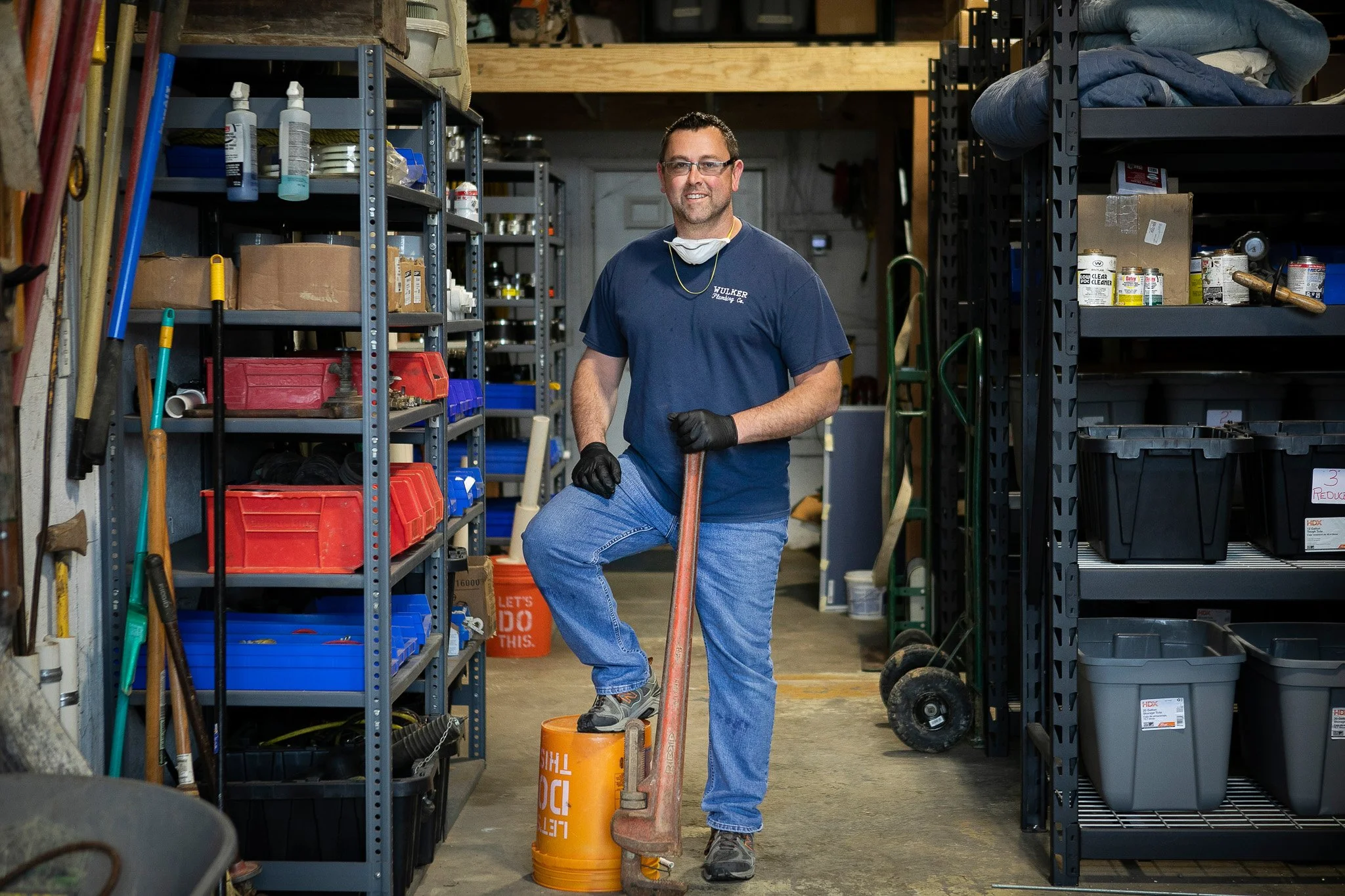 A man is standing in a storage room, holding a crowbar with his foot rested on an orange 20 lb. weight. He is wearing glasses, black gloves, a navy blue T-shirt, and jeans, and is smiling at the camera.