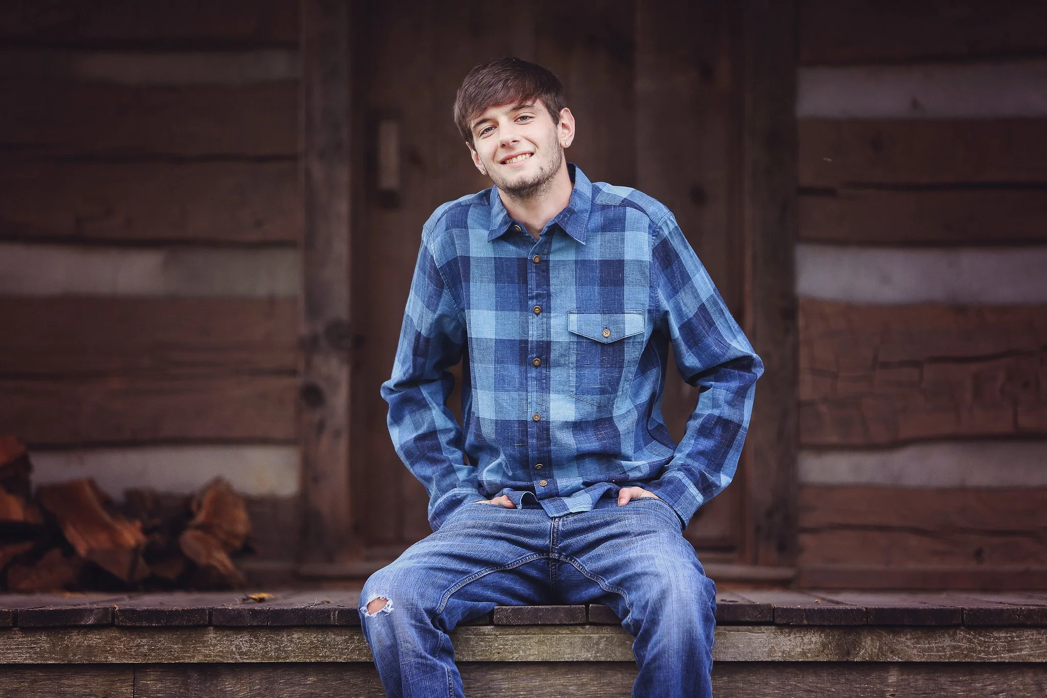 Young man in a blue plaid shirt and ripped jeans sitting on a wooden porch, smiling.