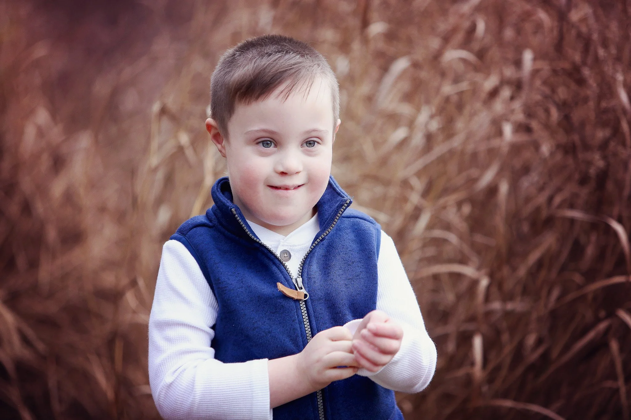 A young boy with short brown hair and light skin, wearing a white long-sleeve shirt and a blue vest, standing outdoors near tall brown grass.