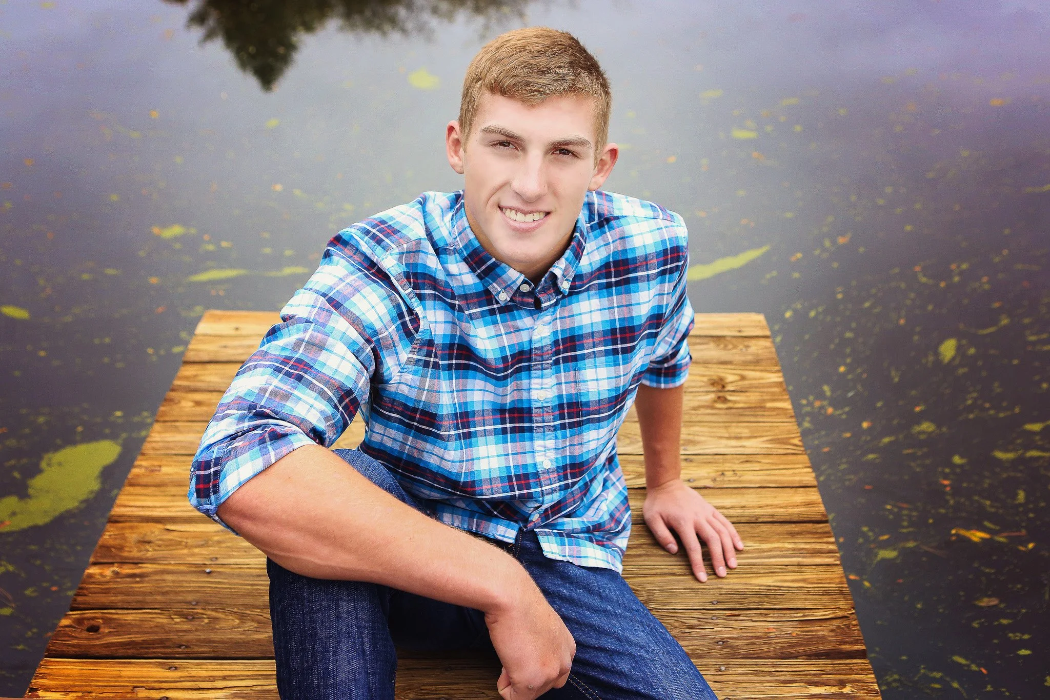 A young man with short blonde hair wearing a blue plaid shirt and jeans, sitting on a wooden dock by the water, smiling at the camera.