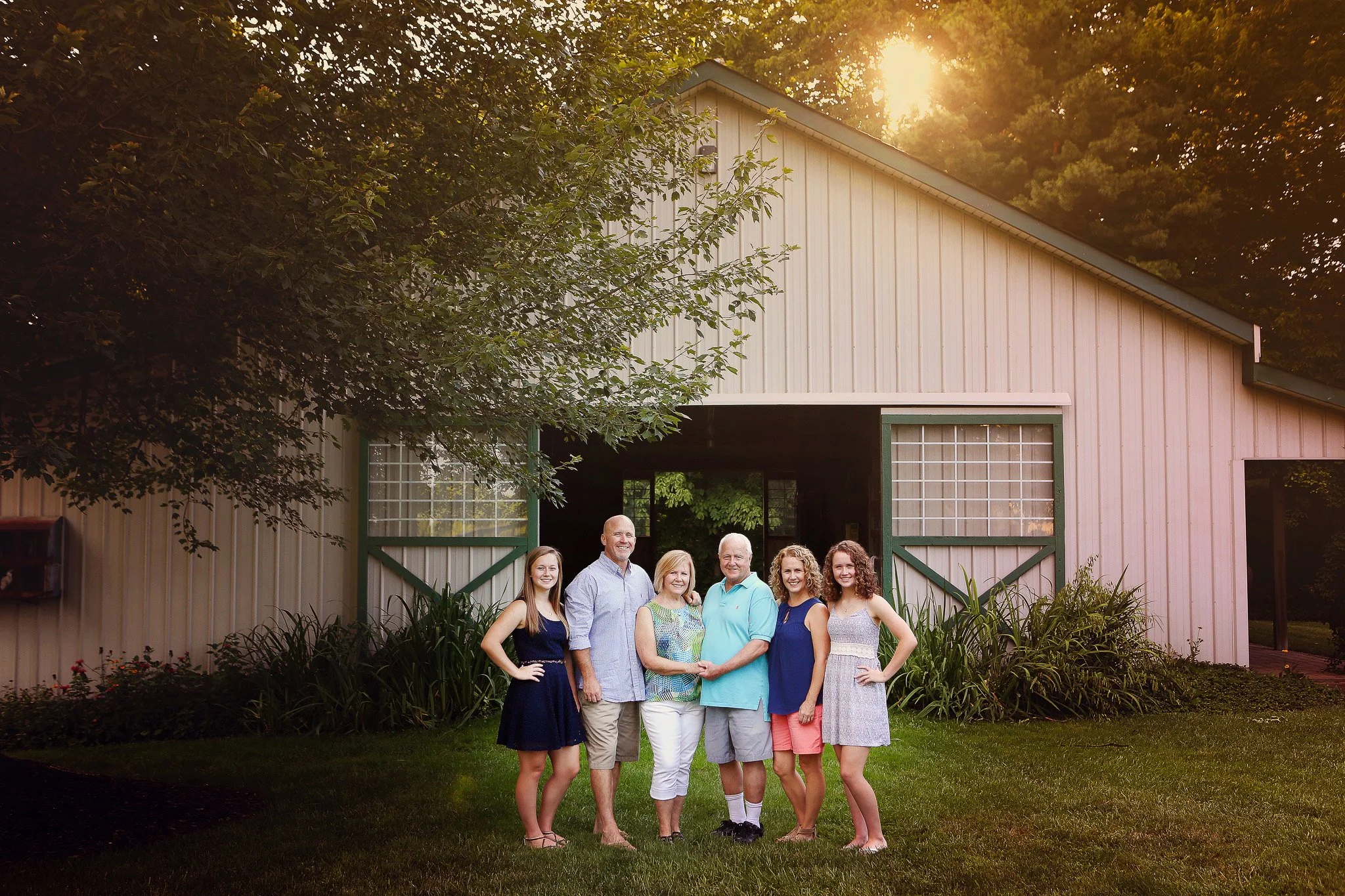 A family of six standing in front of a large barn with open doors, surrounded by greenery, with the sun setting in the background.