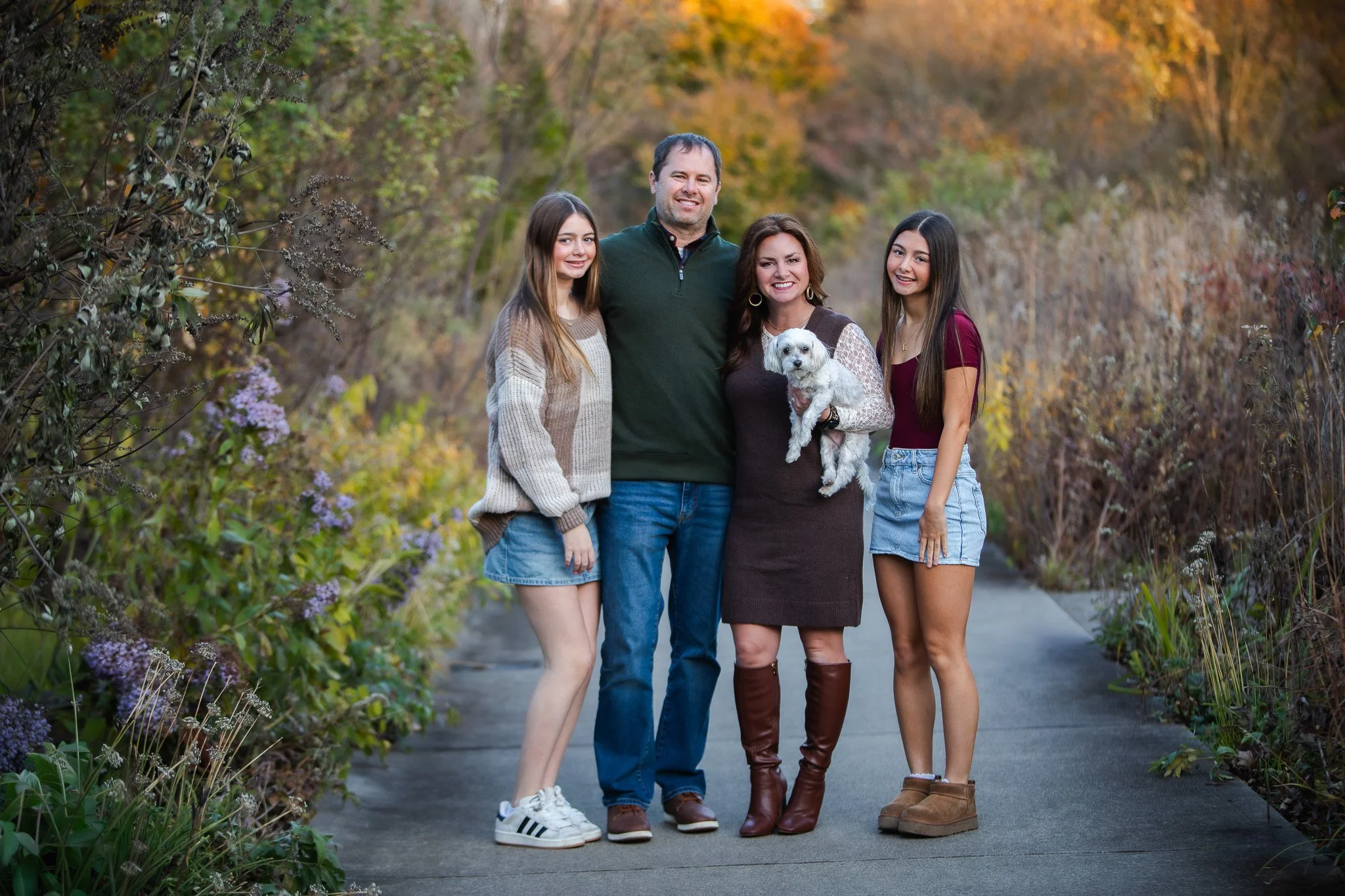 Family of four with two teenage girls and a small white dog standing on a path in a wooded area during fall.