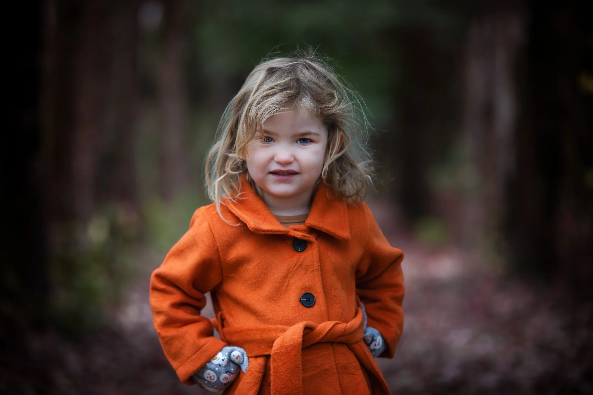 A young girl with blonde hair and blue eyes, wearing an orange coat with a belt, standing outdoors on a forest trail with a blurry background of trees.