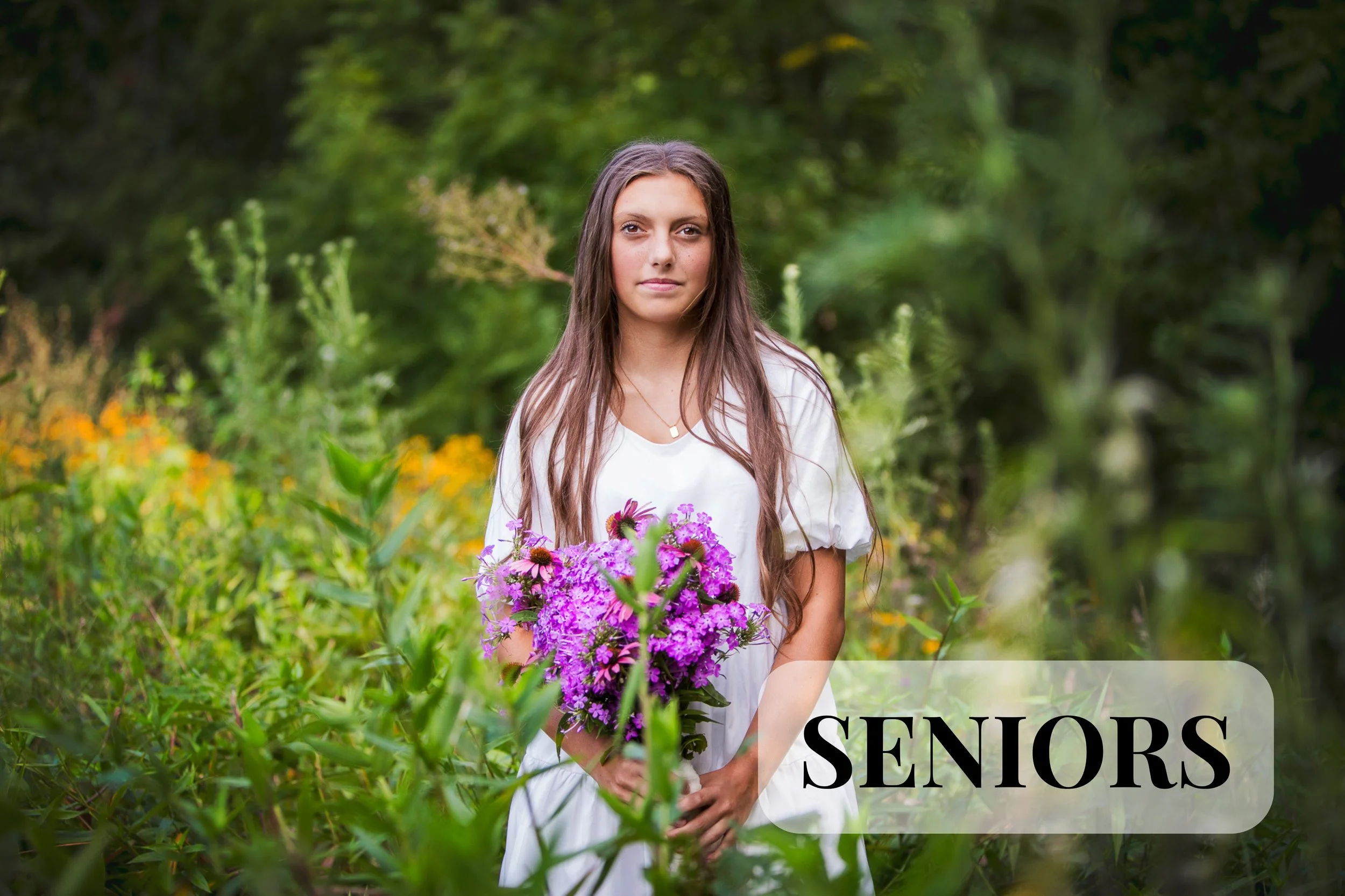 A senior photo session held in Blanchester, Ohio. A teenage girl with long brown hair holds a bouquet of pink and purple flowers outdoors. She is smiling slightly and stands amidst green foliage and yellow flowers, with the word 'SENIORS' partially v