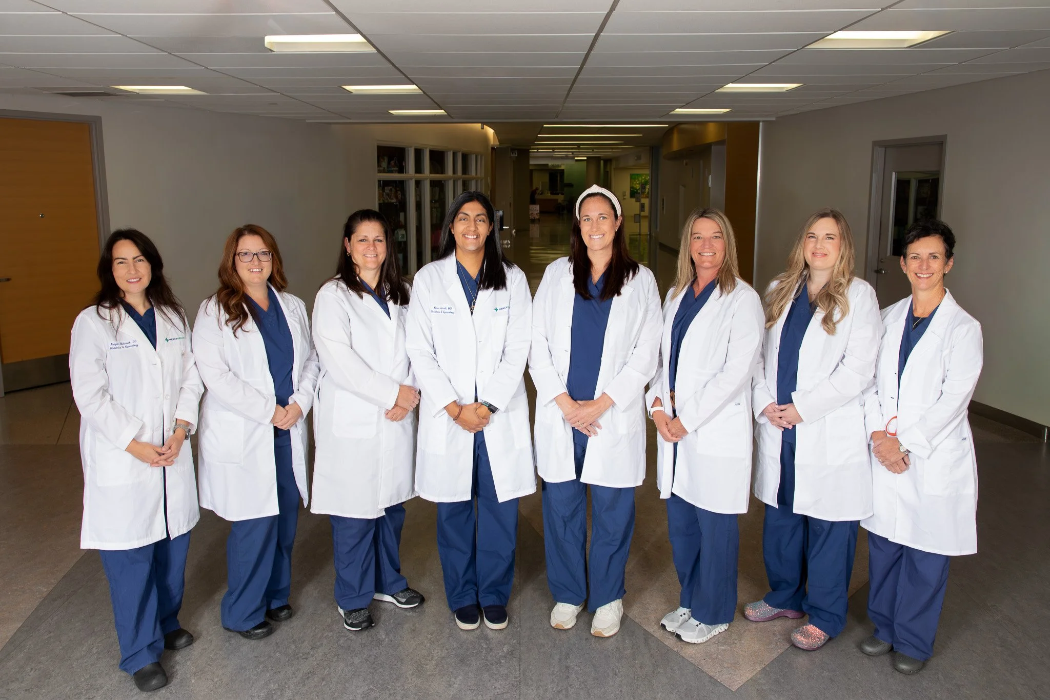 Group of eight women doctors and nurses in white coats and blue scrubs standing in hospital corridor.