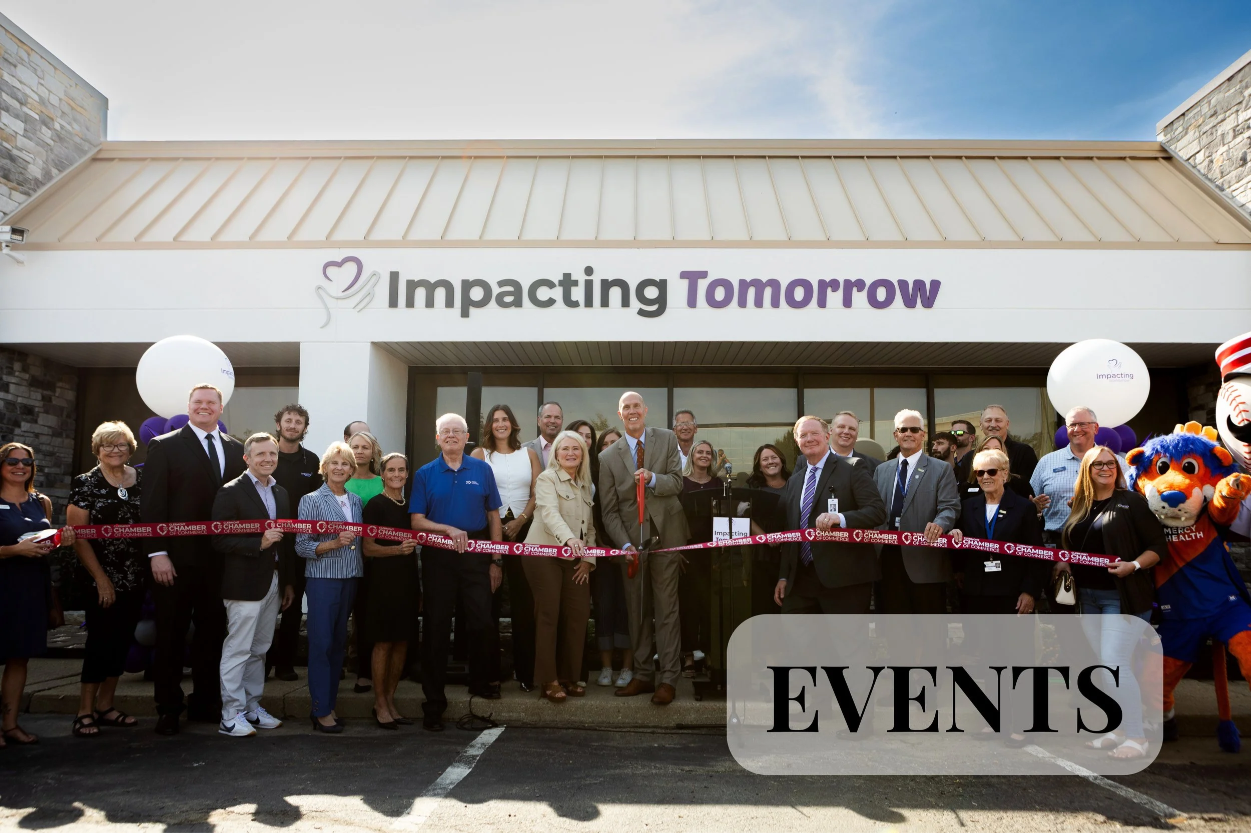 Event coverage of a ribbon cutting for Impacting Tomorrow in Loveland, Ohio. A group of people gathered outside a building with a sign that reads "Impacting Tomorrow" at a ribbon-cutting event, with balloons, a mascot, and a large crowd.