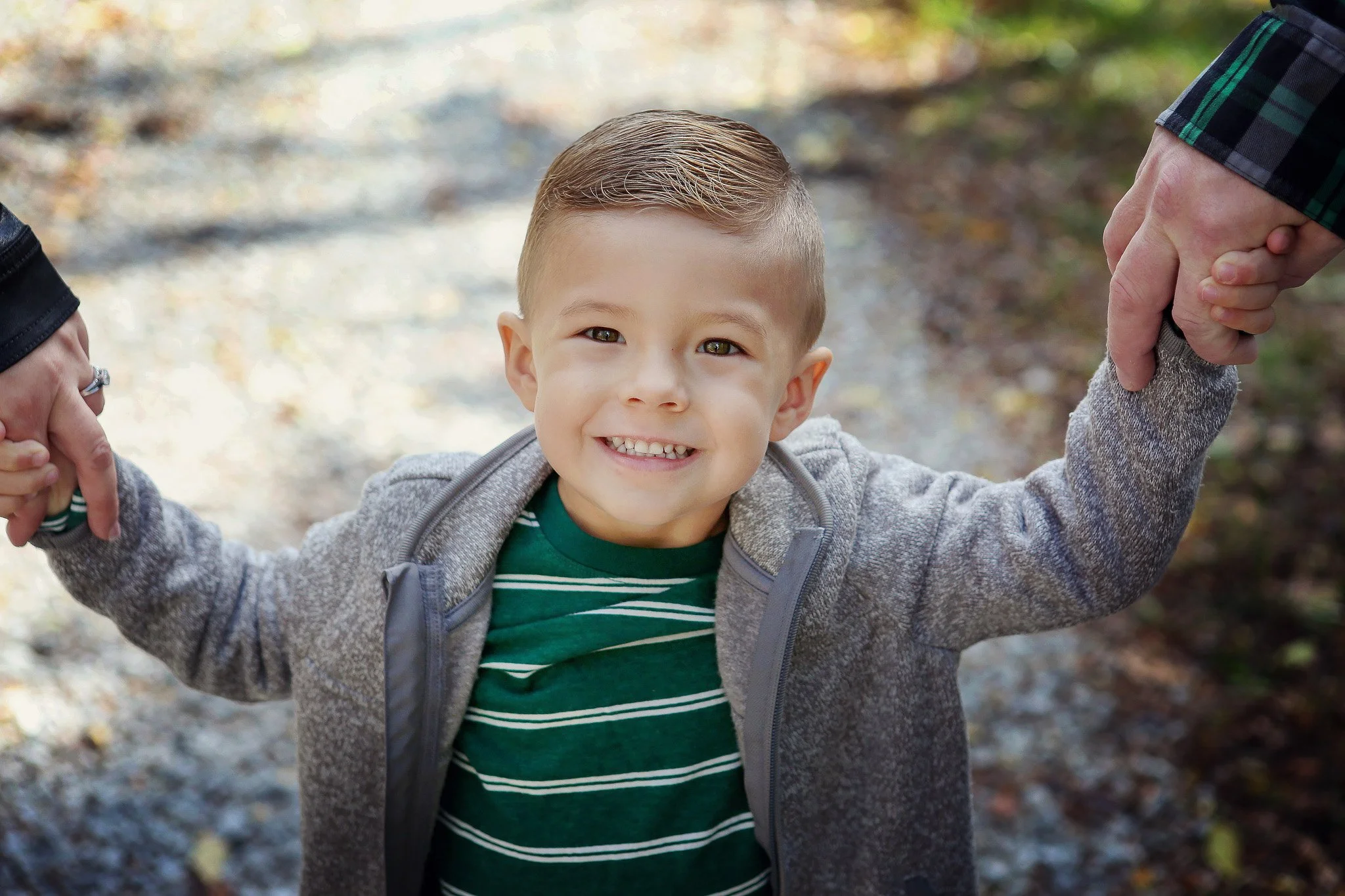 A young boy smiling while holding hands with two adults, outdoors on a trail in autumn, as the adults' hands are visible on each side of the boy.
