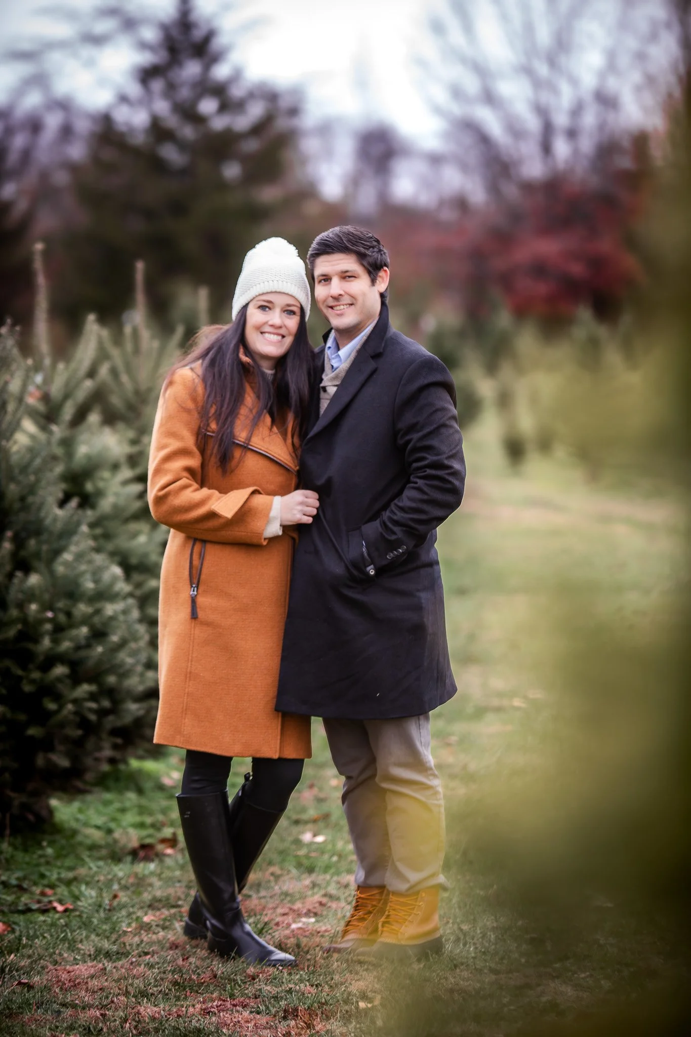 A smiling couple standing outdoors in a park or garden during fall or winter, with trees in the background. The woman is wearing a tan coat, black boots, and a white knit hat. The man is wearing a black coat and tan boots.