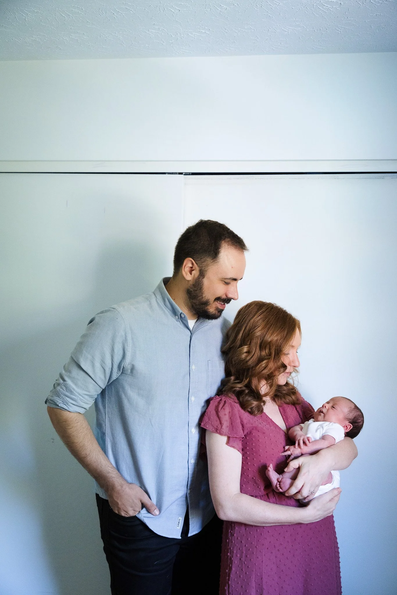 A woman in a maroon dress holding a baby, with a man in a light blue shirt standing beside her, all looking at the baby in her arms in a room with plain white walls.