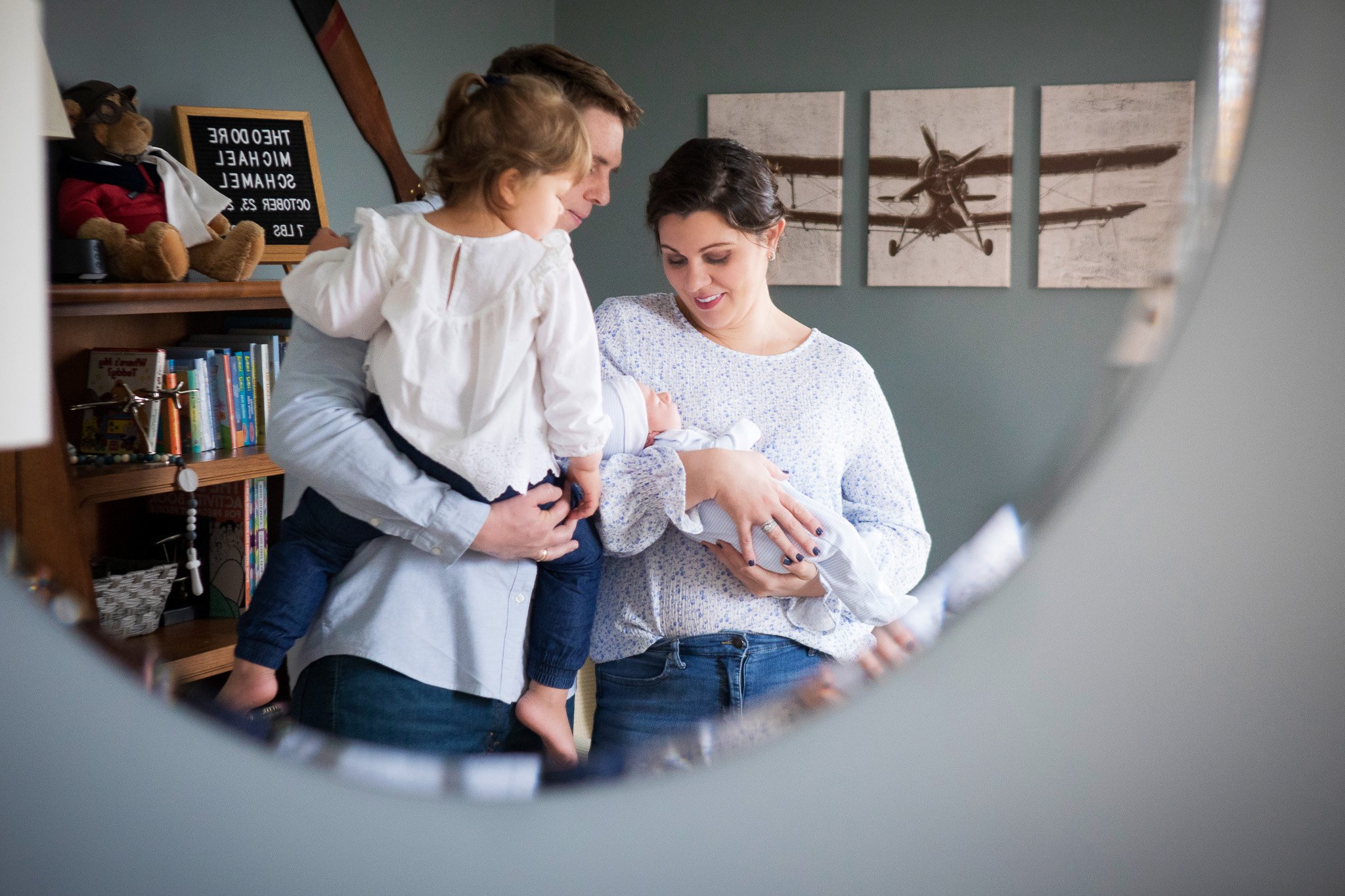 A family gathered around a newborn baby, seen through a circular mirror. The father is holding his young daughter, and the mother is cradling the baby while smiling.