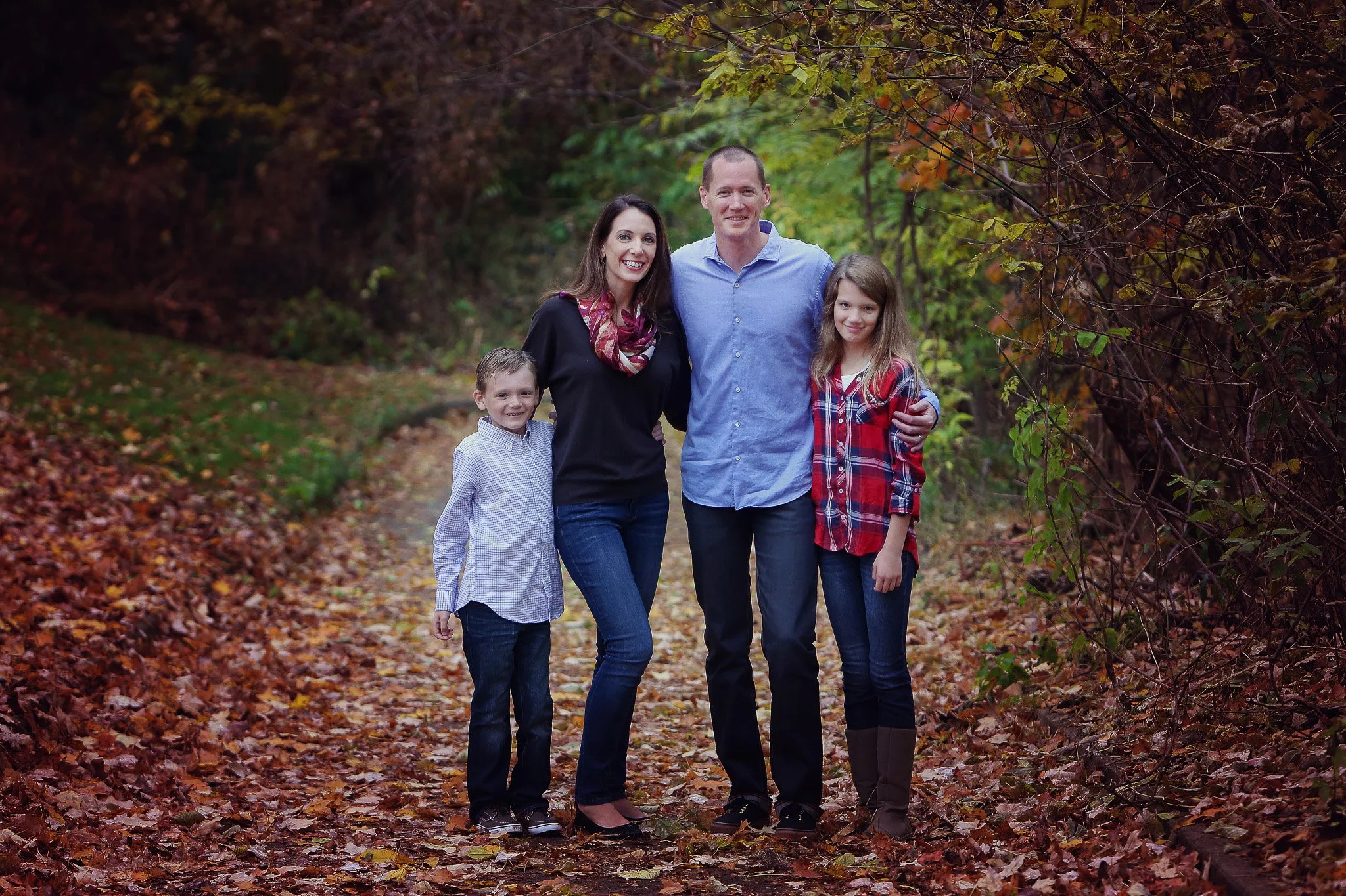 A family of four standing together on a leaf-covered trail in a wooded area during autumn. The mother and father are in the center, with their children on either side. The mother is wearing a black top and jeans, and the father is in a blue shirt and