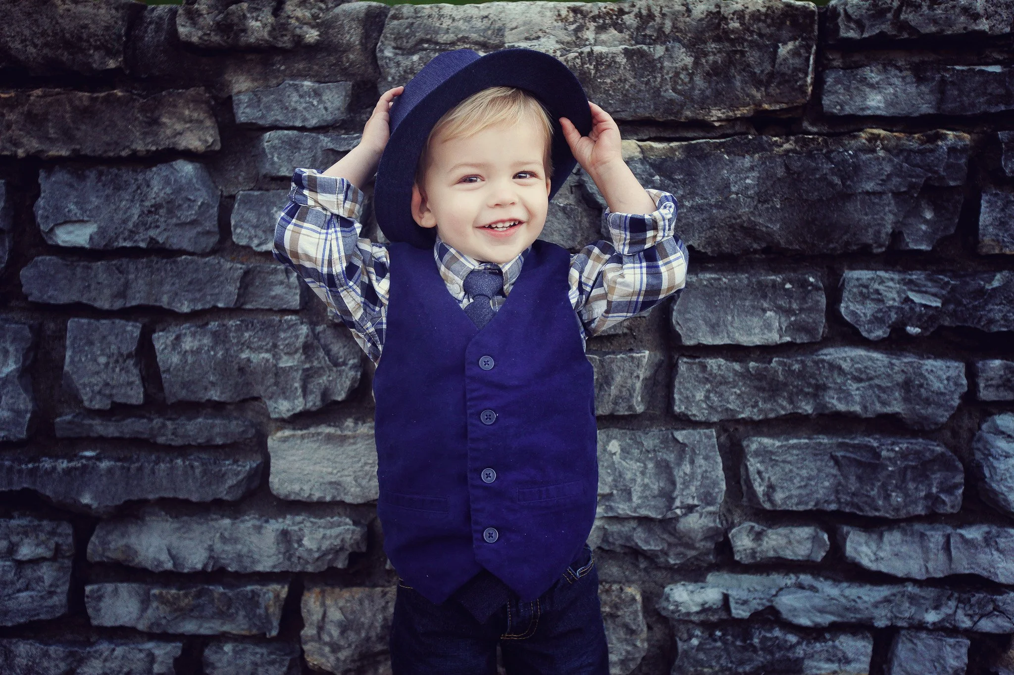 A young boy with blonde hair smiling, wearing a blue vest, checkered shirt, tie, and a dark hat, standing in front of a stone wall.