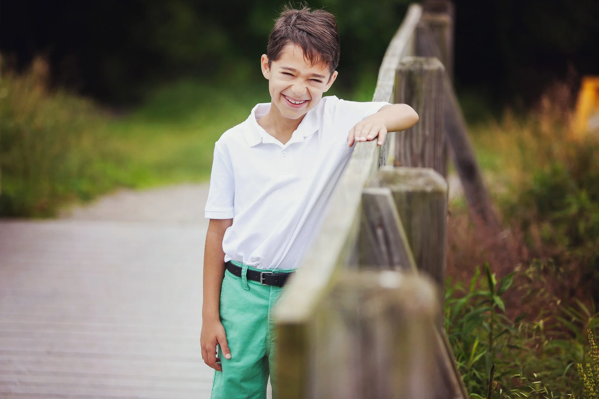 A young boy with short brown hair wearing a white polo shirt and green pants, smiling and leaning on a wooden fence along a paved path in a park or natural outdoor setting.