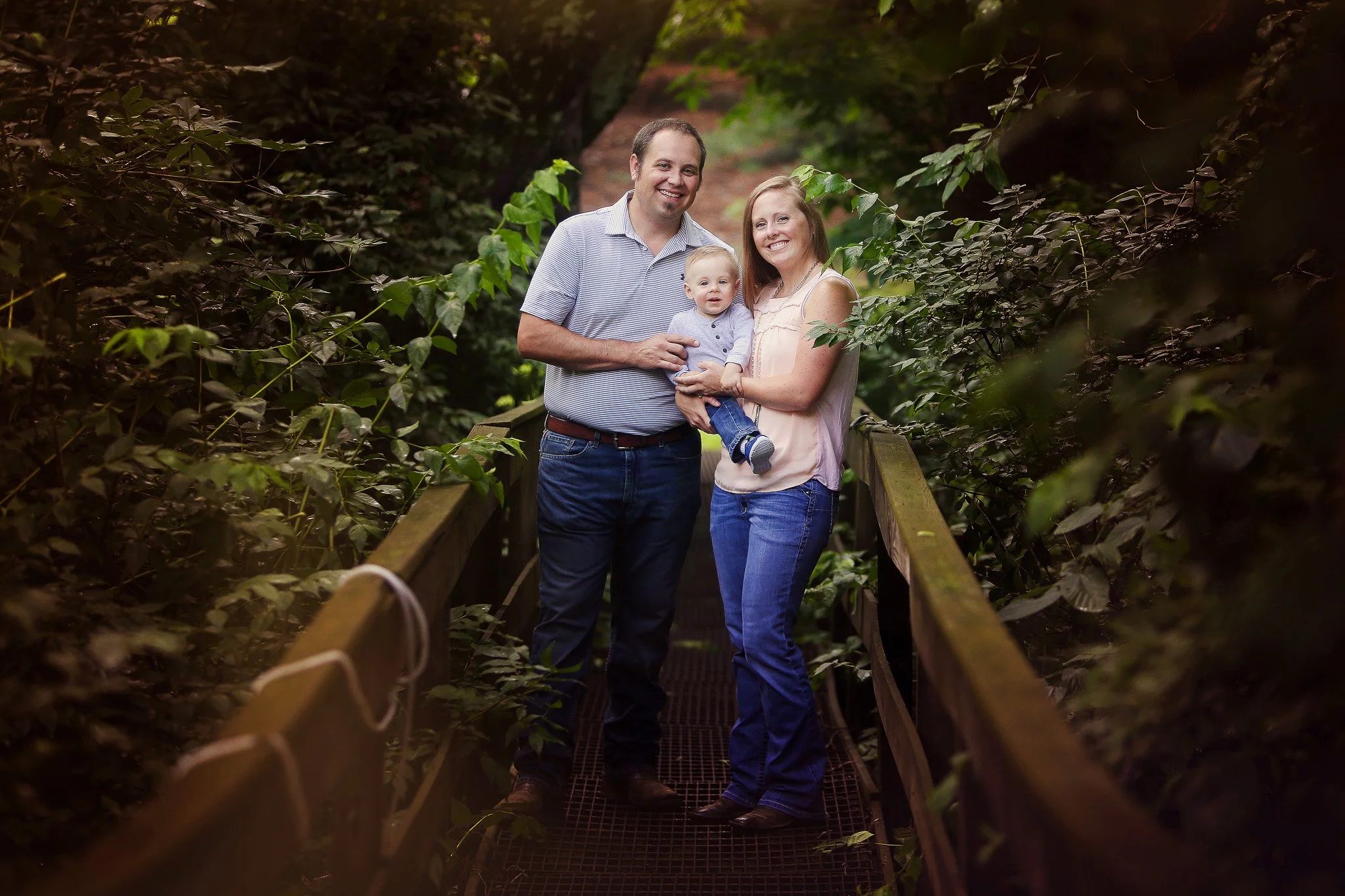 Family of three (father, mother, and young child) standing on a wooden bridge surrounded by lush greenery.