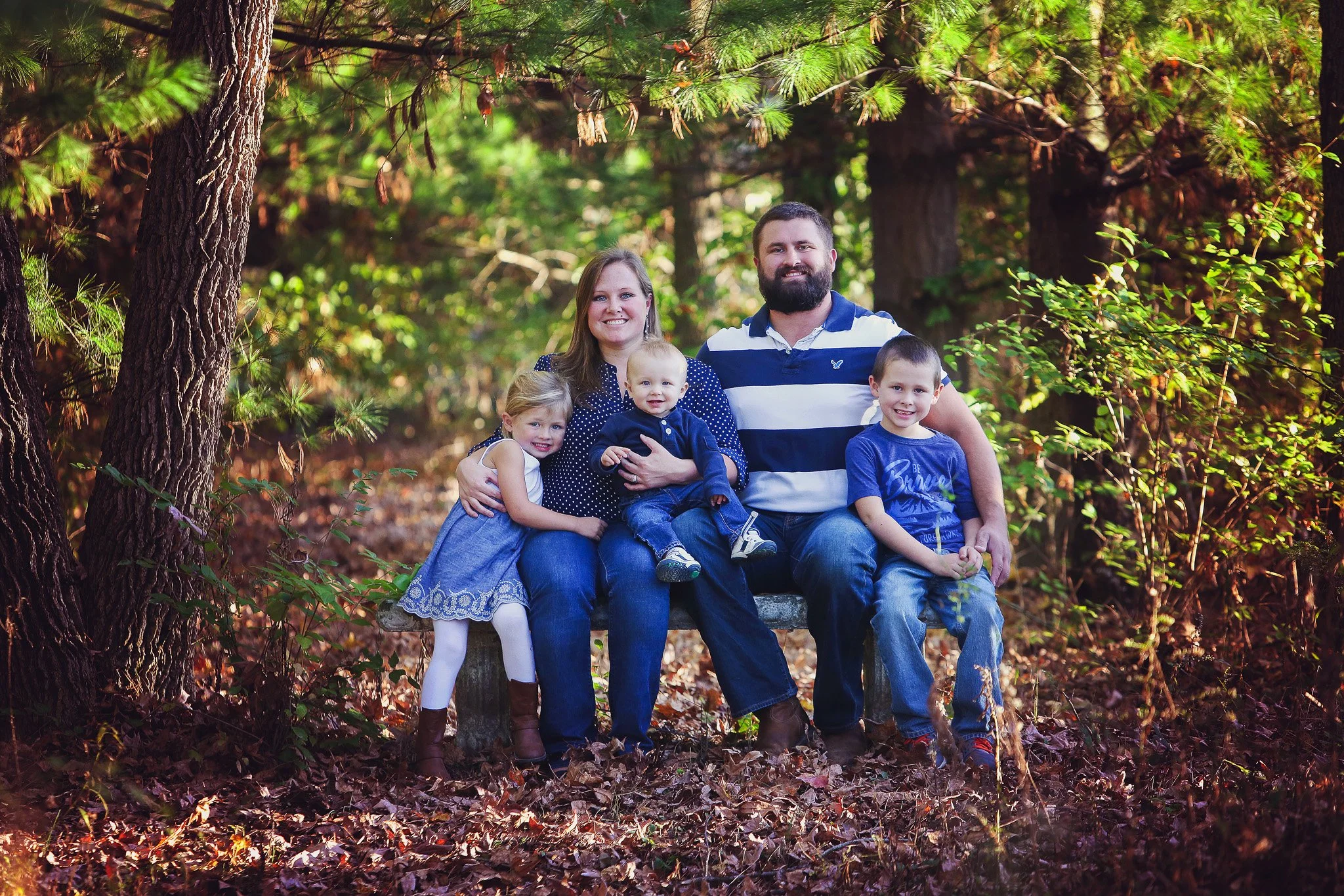 Family of five sitting on a park bench surrounded by trees and fallen leaves, smiling at the camera.
