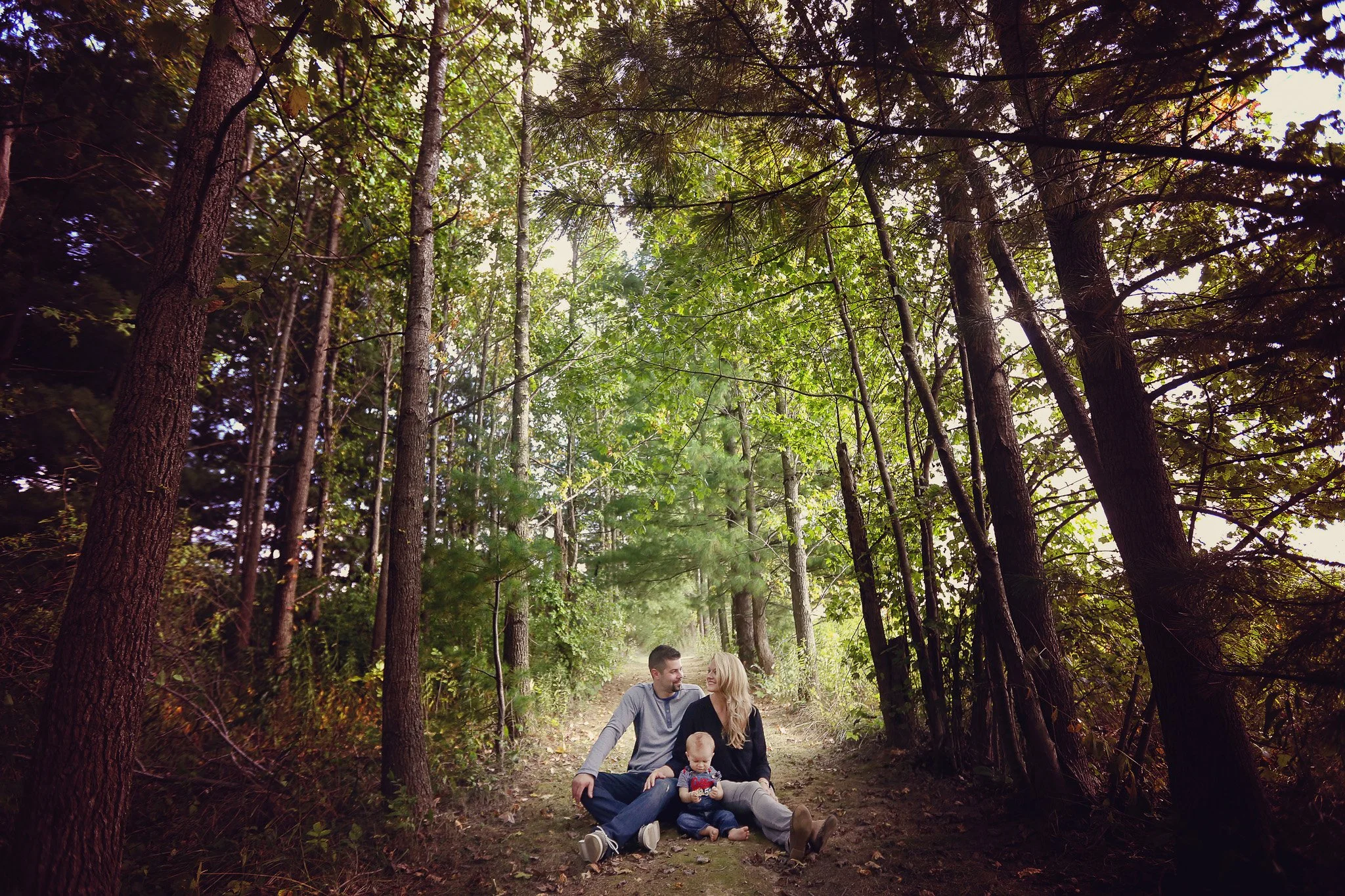 A family of three sitting on a forest trail, surrounded by tall trees and green foliage, enjoying a moment together.