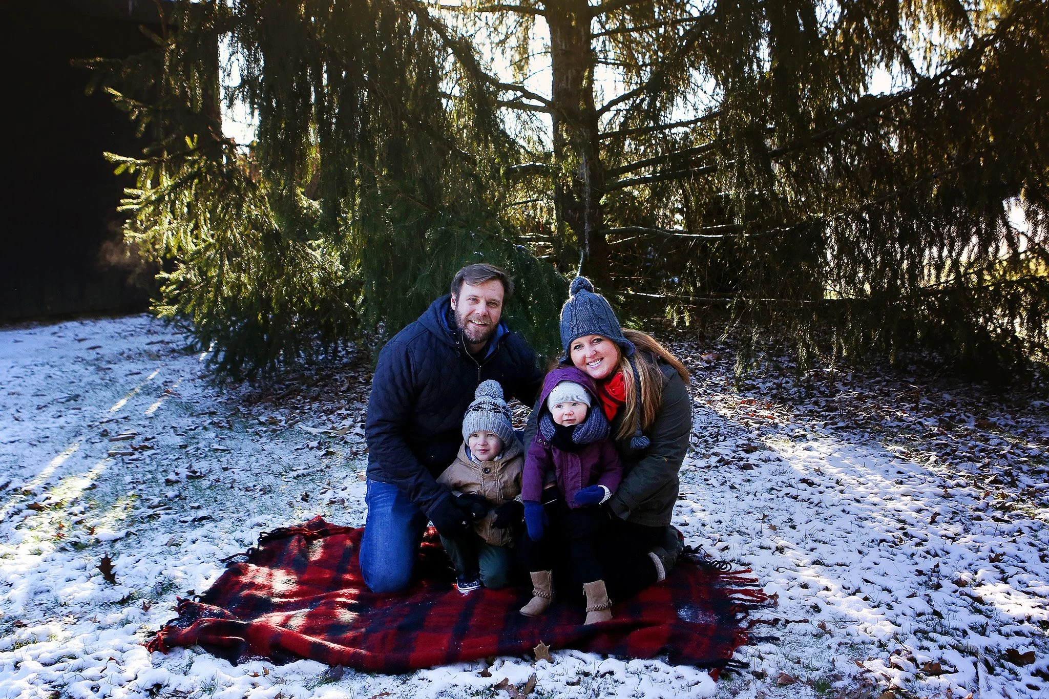 A family of four sitting on a red and black plaid blanket outdoors in a snow-covered forest, dressed warmly with hats and gloves. The parents are smiling, with the mother wearing a gray knit hat and the father in a dark jacket. The children are stand