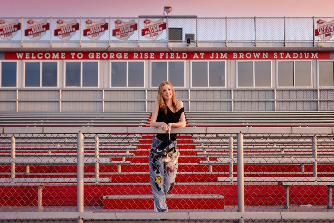 A young woman standing on red stadium bleachers at Goshen High School in front of a chain-link fence at George Rise Field at Jim Brown Stadium, with banners hanging above and a pink sky in the background.