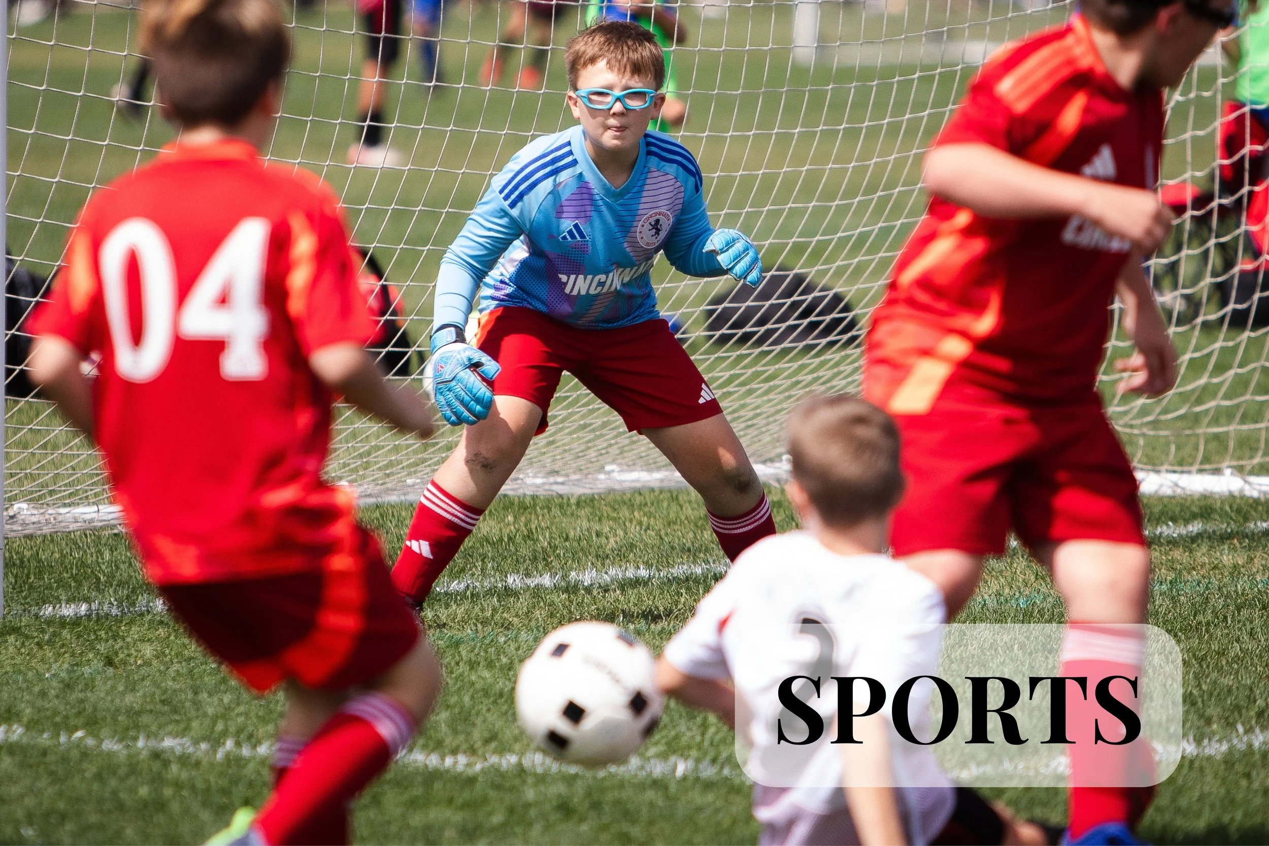 Game Day photos from a soccer team in Loveland, Ohio. Young boys playing soccer on a grassy field, with a goalkeeper in a blue jersey and goggles, ready to block a shot, surrounded by other kids in red uniforms, with a soccer ball near their feet and