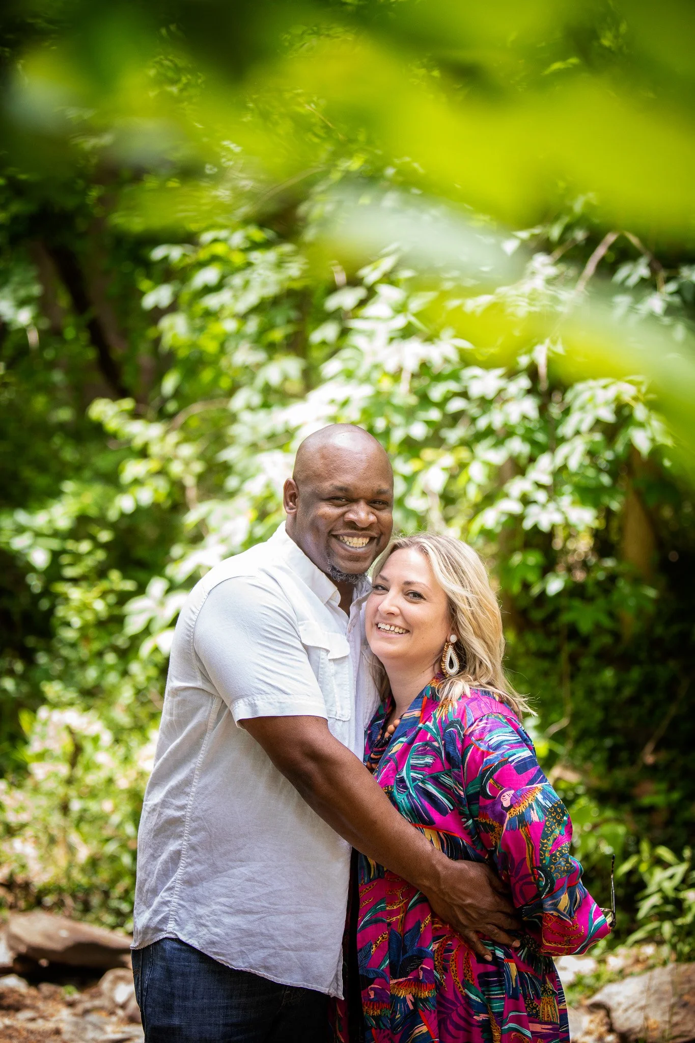 A smiling couple hugging in a lush green outdoor setting.