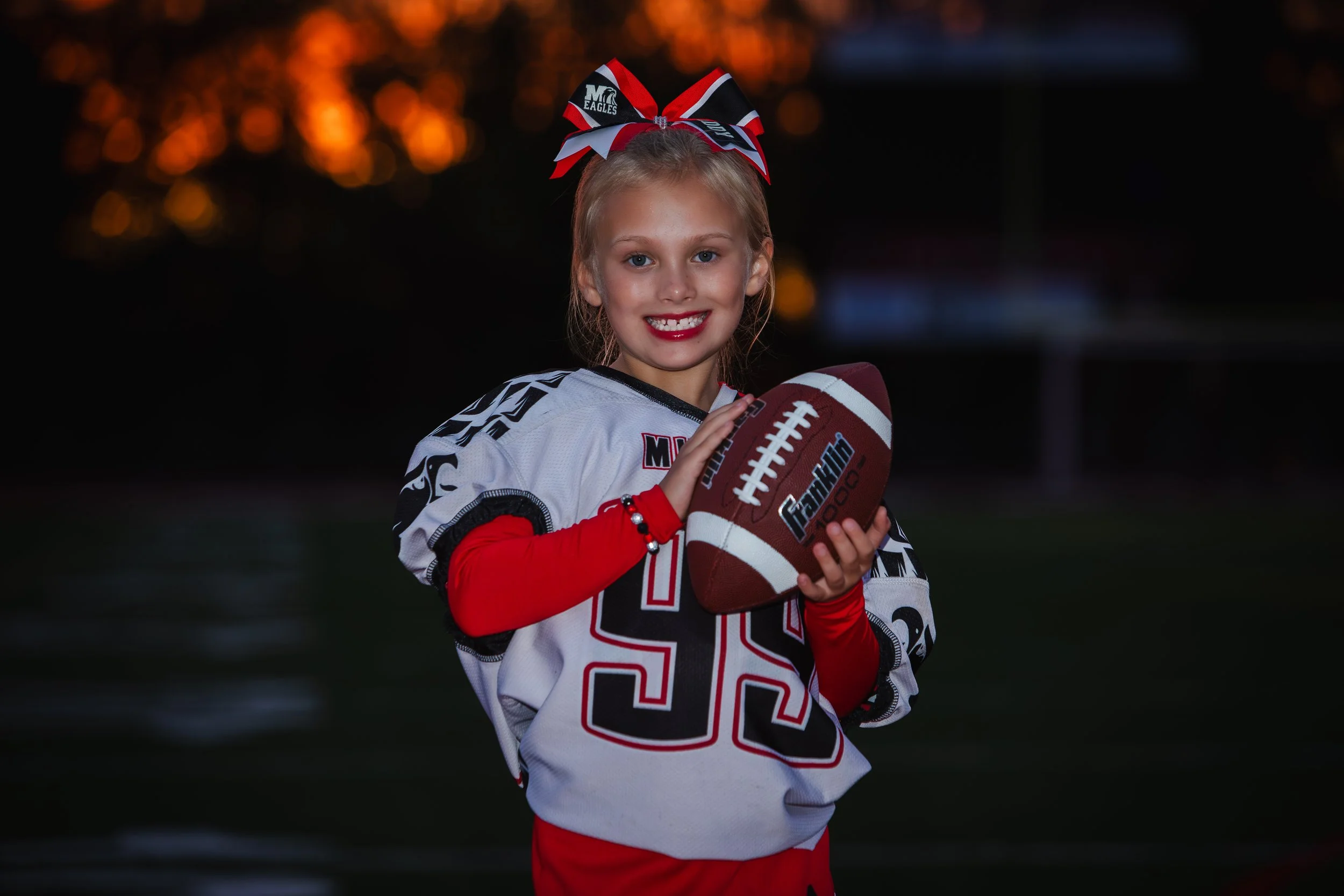 Young girl in football uniform with red, white, and black bow in her hair, holding a football, smiling on football field during sunset.
