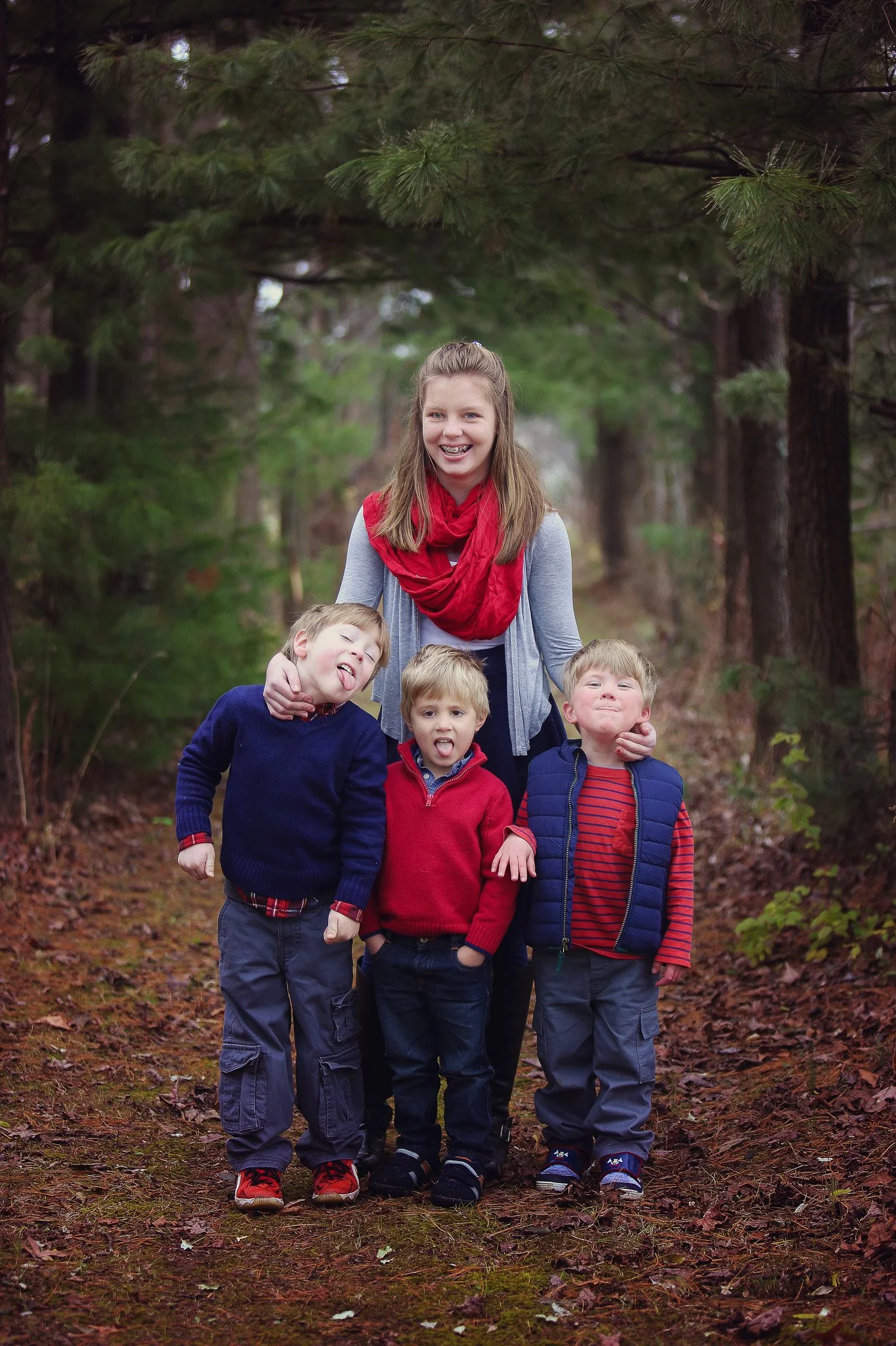 A young woman with long hair smiling and wearing a red scarf, gray cardigan, and black pants, standing in a forest with three young boys in front of her. The boys are dressed in casual clothing, with two wearing red sweaters and one in a navy vest, s