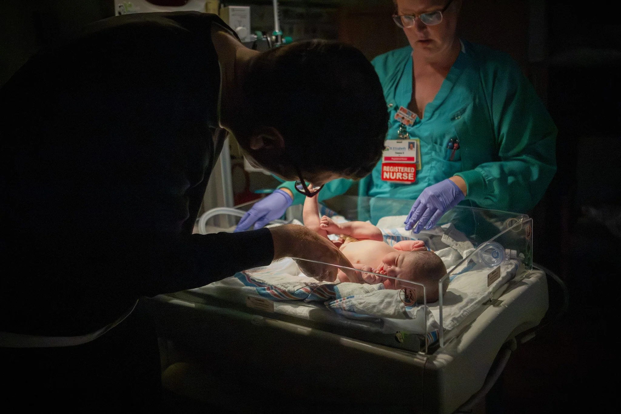 A newborn baby being examined by two healthcare professionals in a dimly lit hospital room. One professional is wearing a teal scrubs with a 'Registered Nurse' badge and purple gloves, while the other is leaning over the baby, wearing glasses and dar