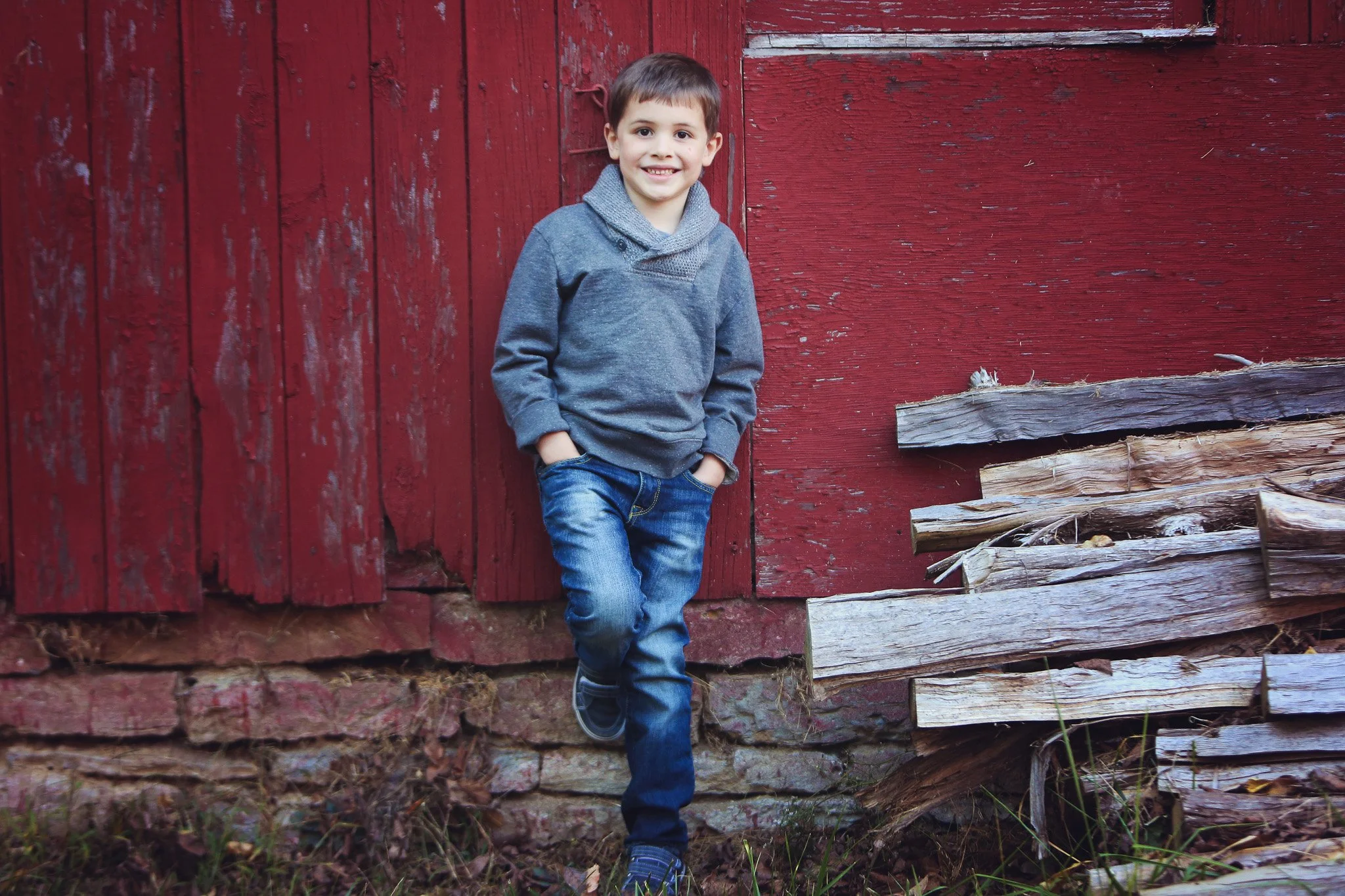 A young boy leaning against a weathered red barn, smiling with hands in pockets, wearing a gray hoodie, blue jeans, and sneakers.