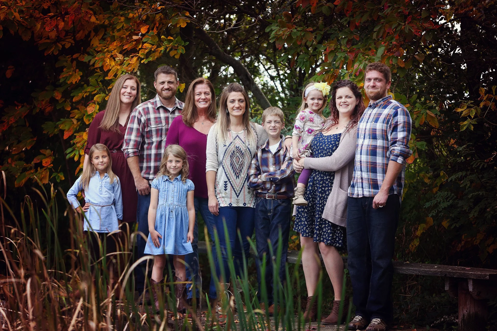 Family group photo outdoors in fall, with twelve people including children, standing on a wooden bridge surrounded by trees with autumn leaves.