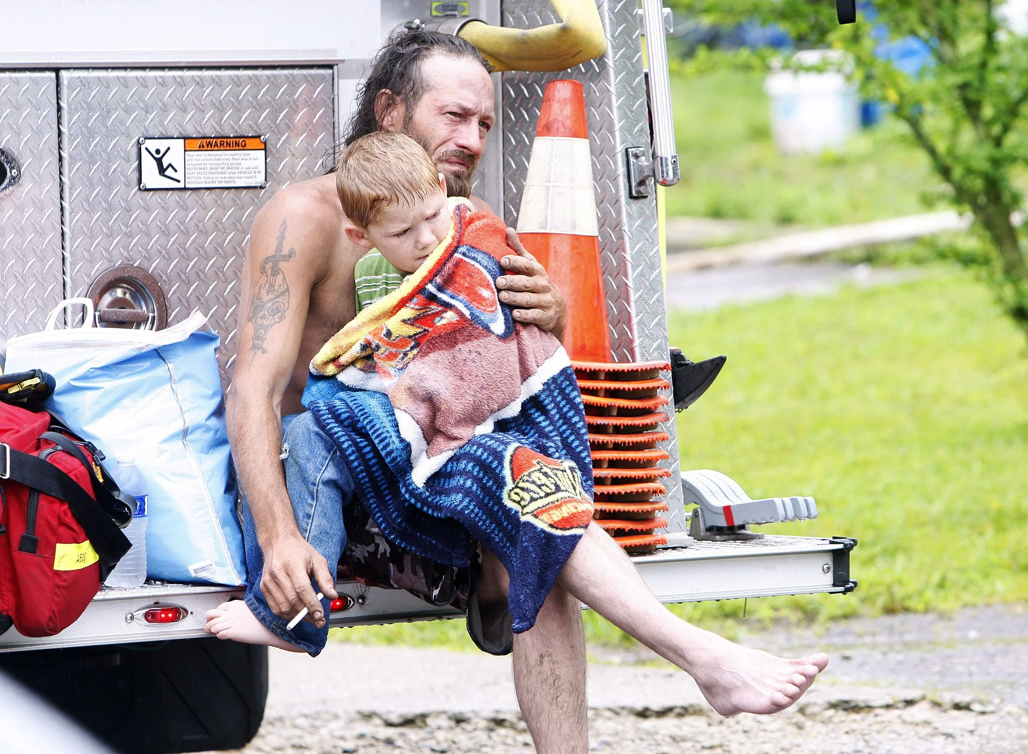 A shirtless man with tattoos sitting on the back of a fire truck, holding a young boy wrapped in a towel, both appearing tired or upset. There are traffic cones and emergency equipment around them, with a grassy outdoor area in the background.
