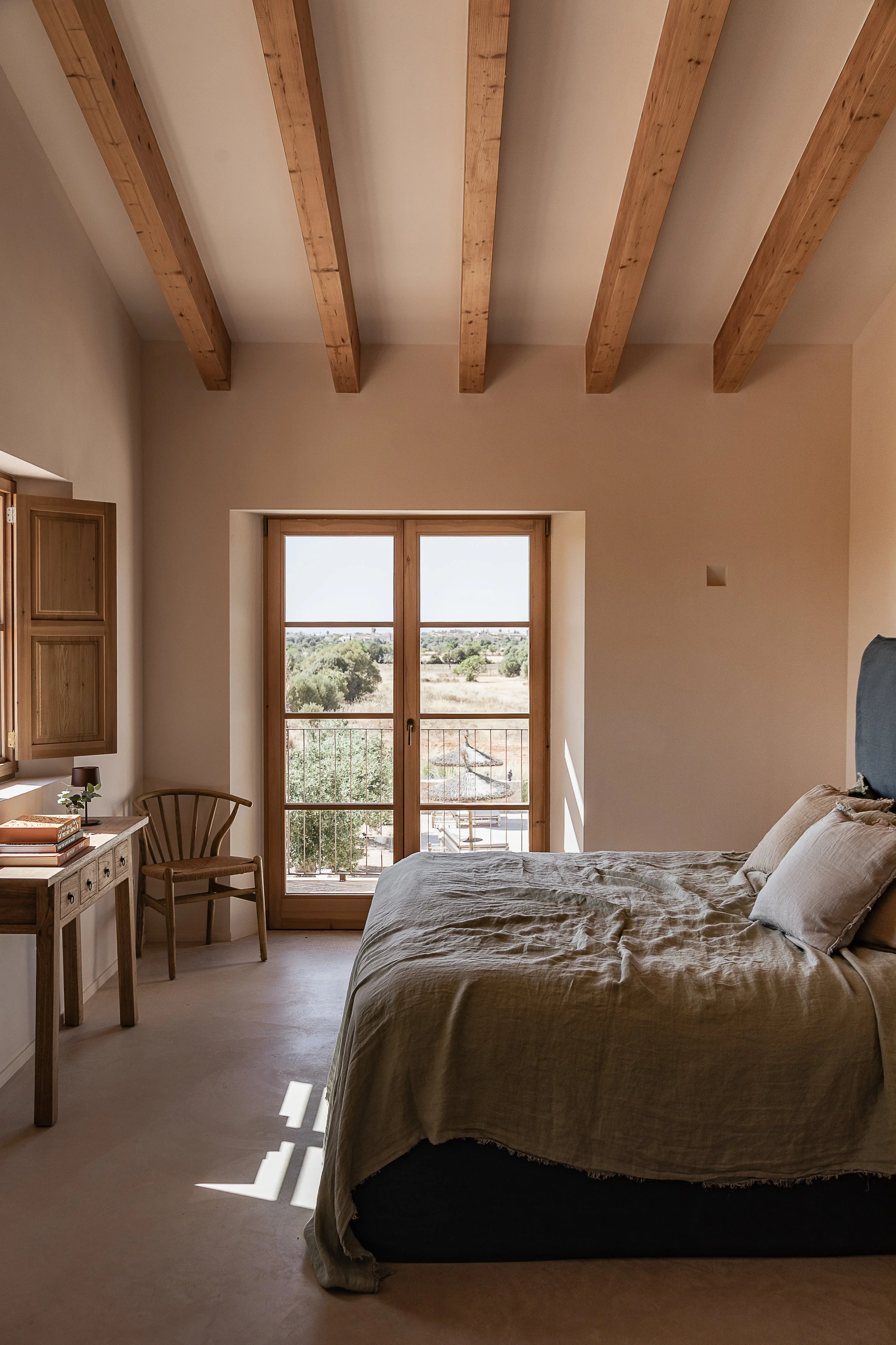 Bedroom with exposed timber beams, oak-framed doors opening to the Mallorcan countryside and seamless poured concrete flooring in a Mediterranean home.